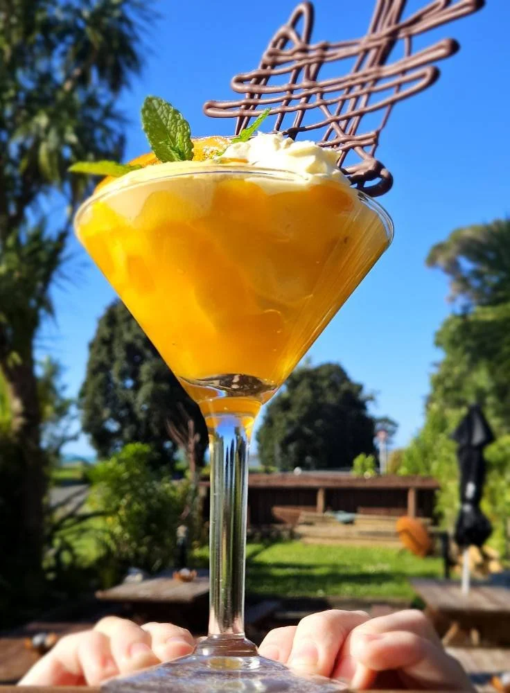 Close-up of a yellow-orange cocktail in a martini glass, garnished with a mint leaf and a drizzle chocolate decoration, held outdoors against a blue sky and green trees.