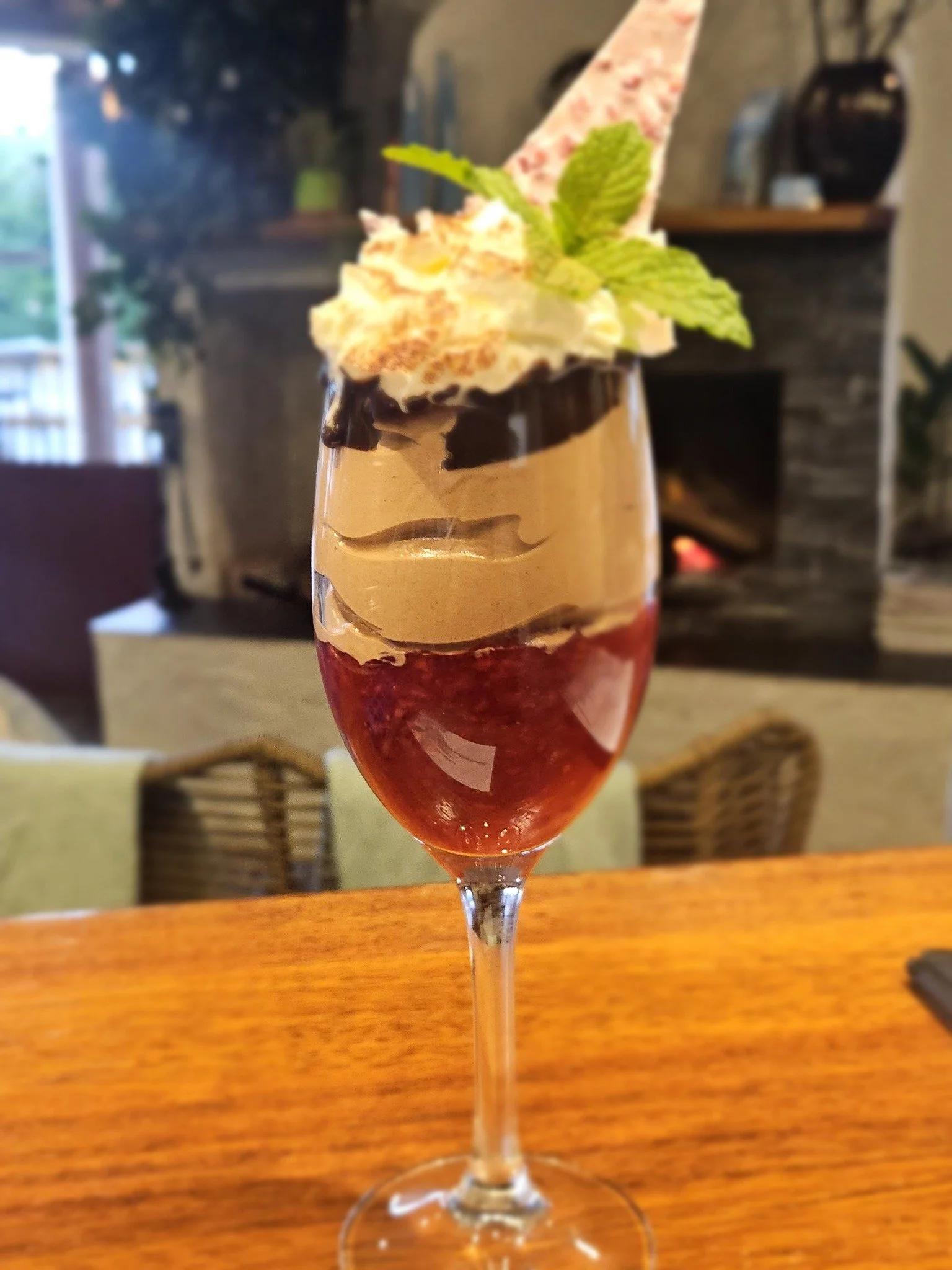 A layered dessert in a tall glass with red, orange, and beige layers, topped with whipped cream, a mint leaf, and a decorative cookie piece. The dessert is on a wooden table in a cozy restaurant setting.