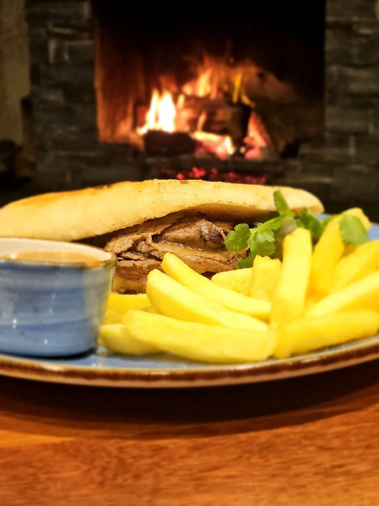 Plate with French fries, a sandwich with grilled meat, and garnished with cilantro, placed in front of a fireplace with burning logs.