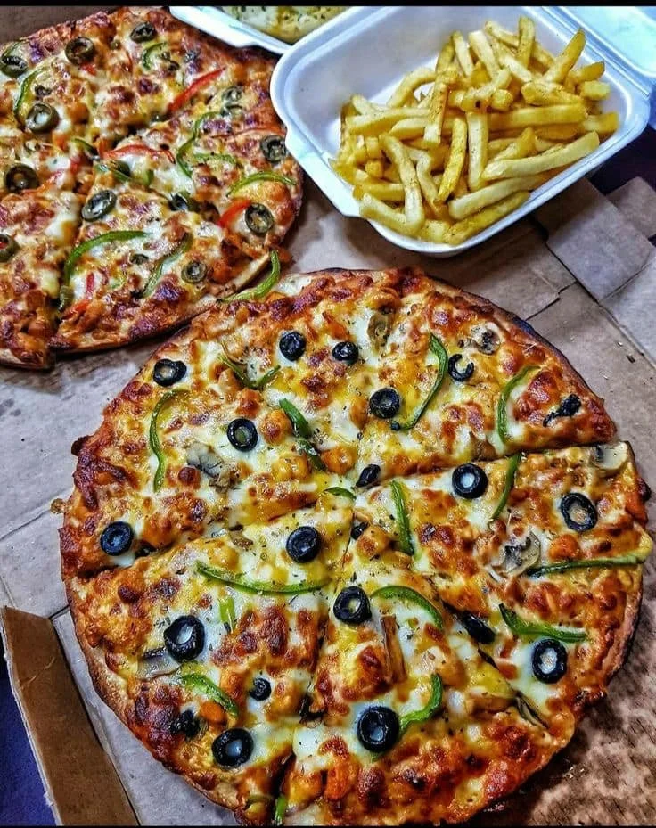 A pizza topped with black olives, green bell peppers, and cheese, along with a slice of another pizza with similar toppings, and a side of French fries in a container.