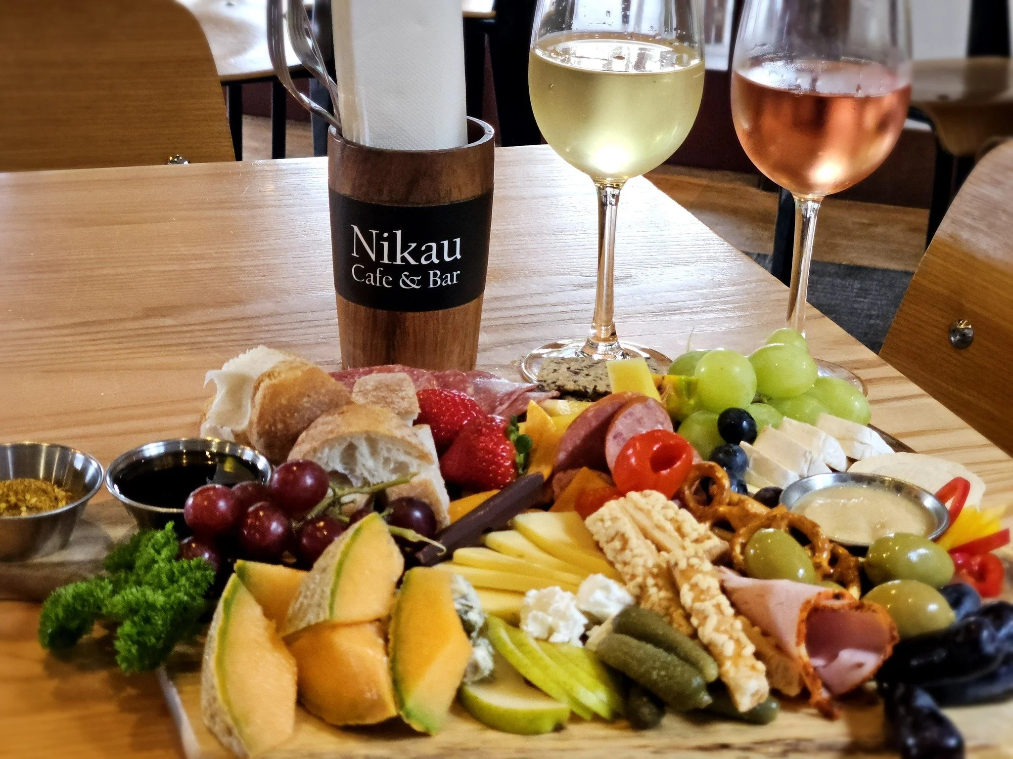 A cheese and fruit platter with grapes, sliced melon, and assorted cheeses, with two glasses of white and rosé wine on a wooden table at a restaurant named Nikau Cafe & Bar.