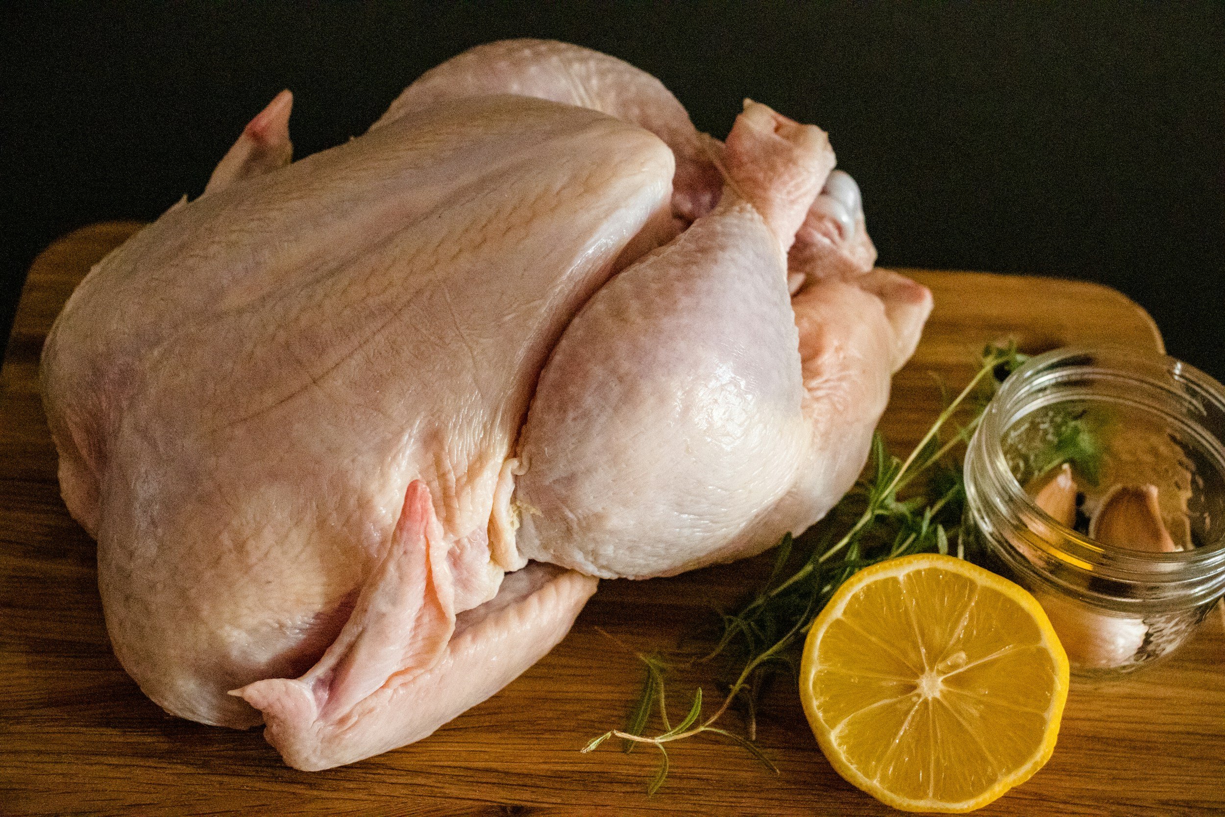 Raw whole chicken on a wooden cutting board, garnished with lemon slices and fresh herbs, with a small jar of marinade or sauce nearby.