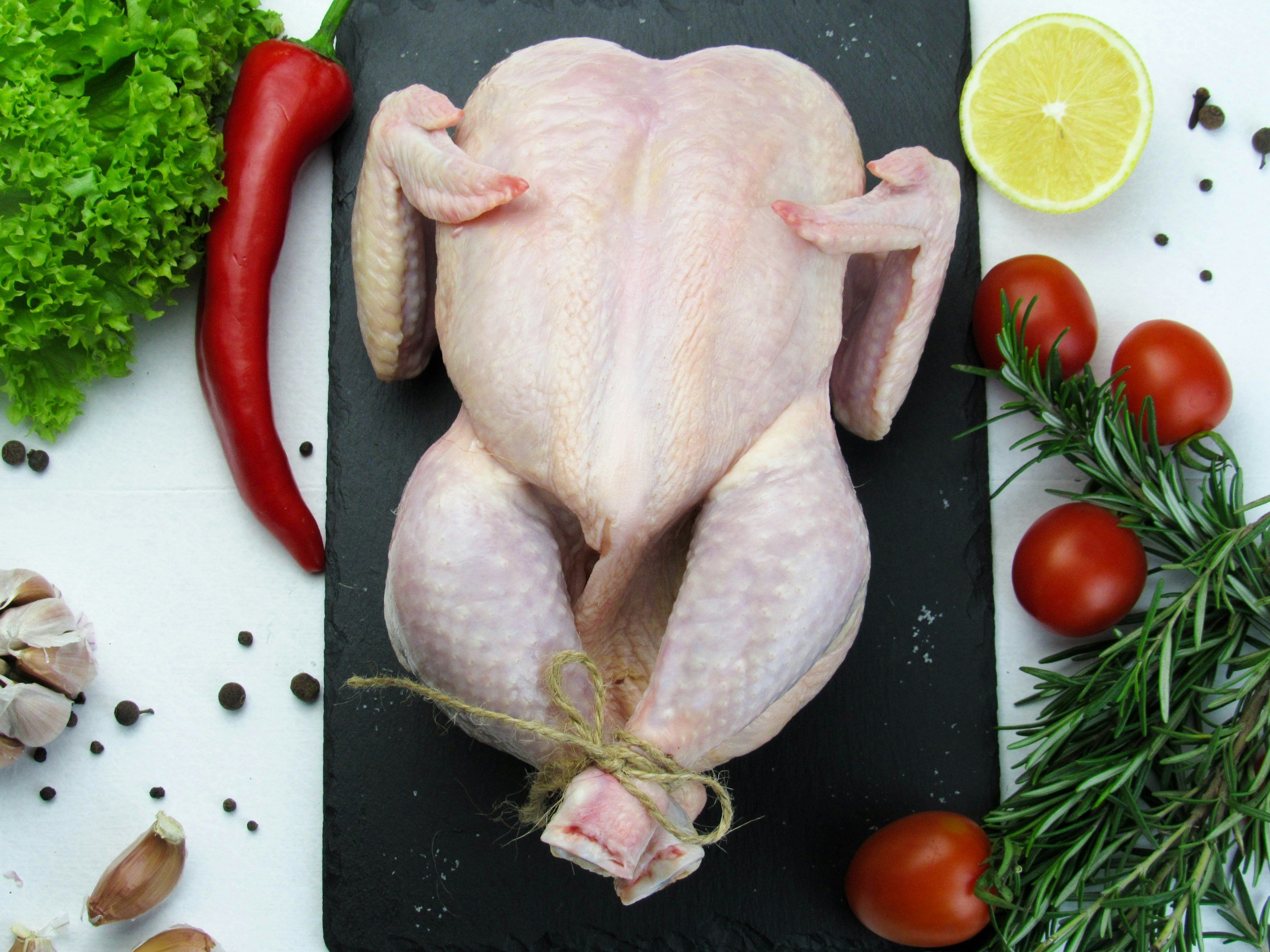 Raw whole chicken placed on a black cutting board surrounded by fresh vegetables and herbs, including lettuce, red chili, garlic cloves, cherry tomatoes, lemon half, and sprigs of rosemary and peppercorns.