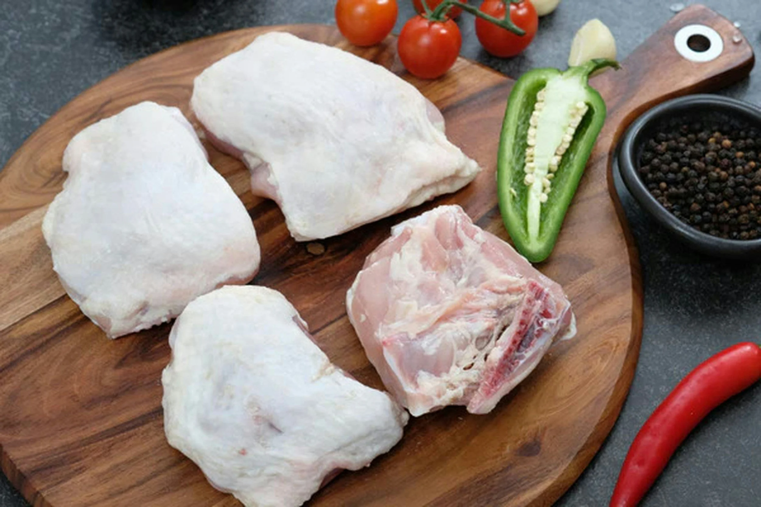 Raw chicken pieces, cherry tomatoes on the vine, a halved green bell pepper, black peppercorns in a bowl, and a piece of garlic on a wooden cutting board.