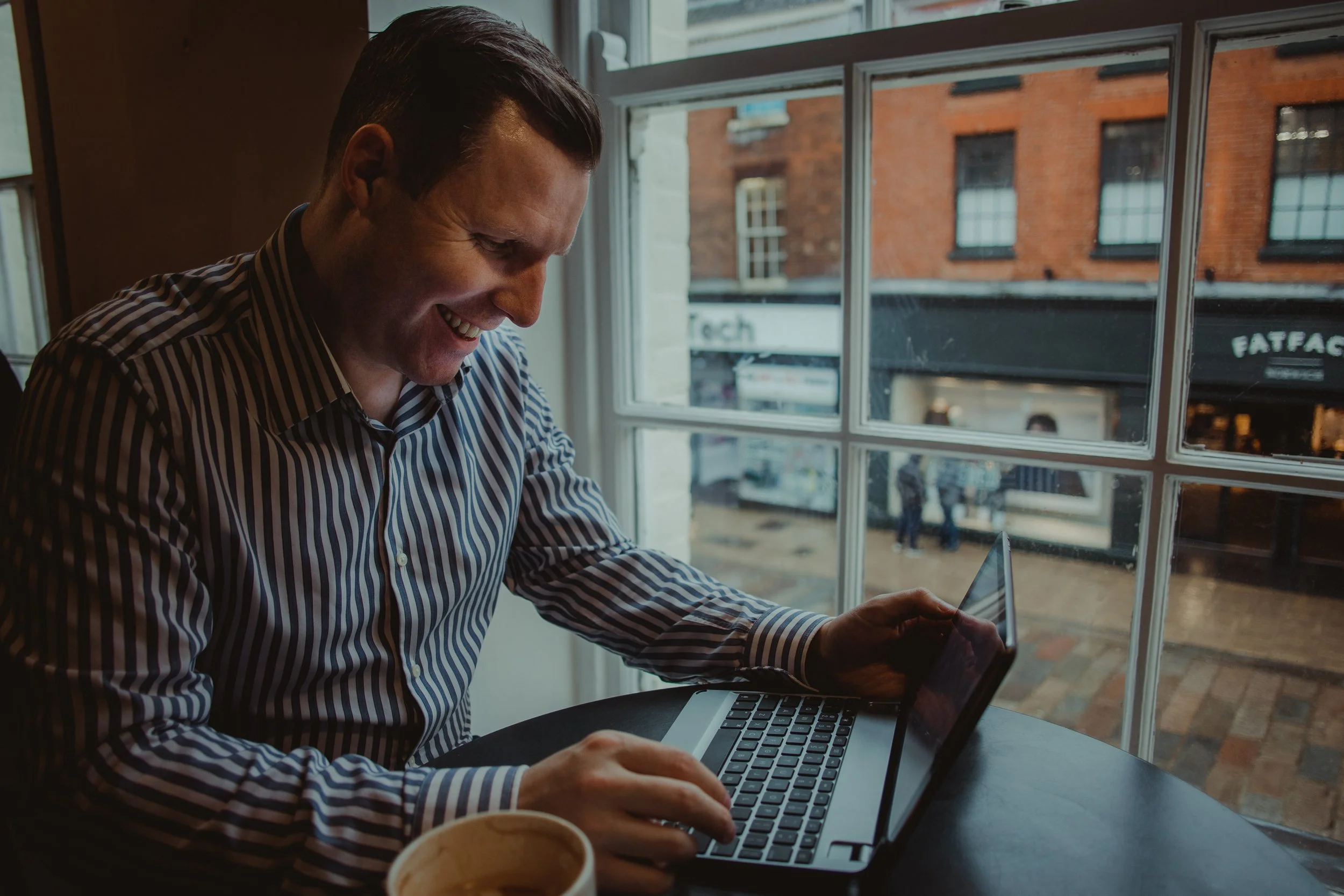 Man in striped shirt working on a laptop at a cafe near a window with a view of brick buildings outside.