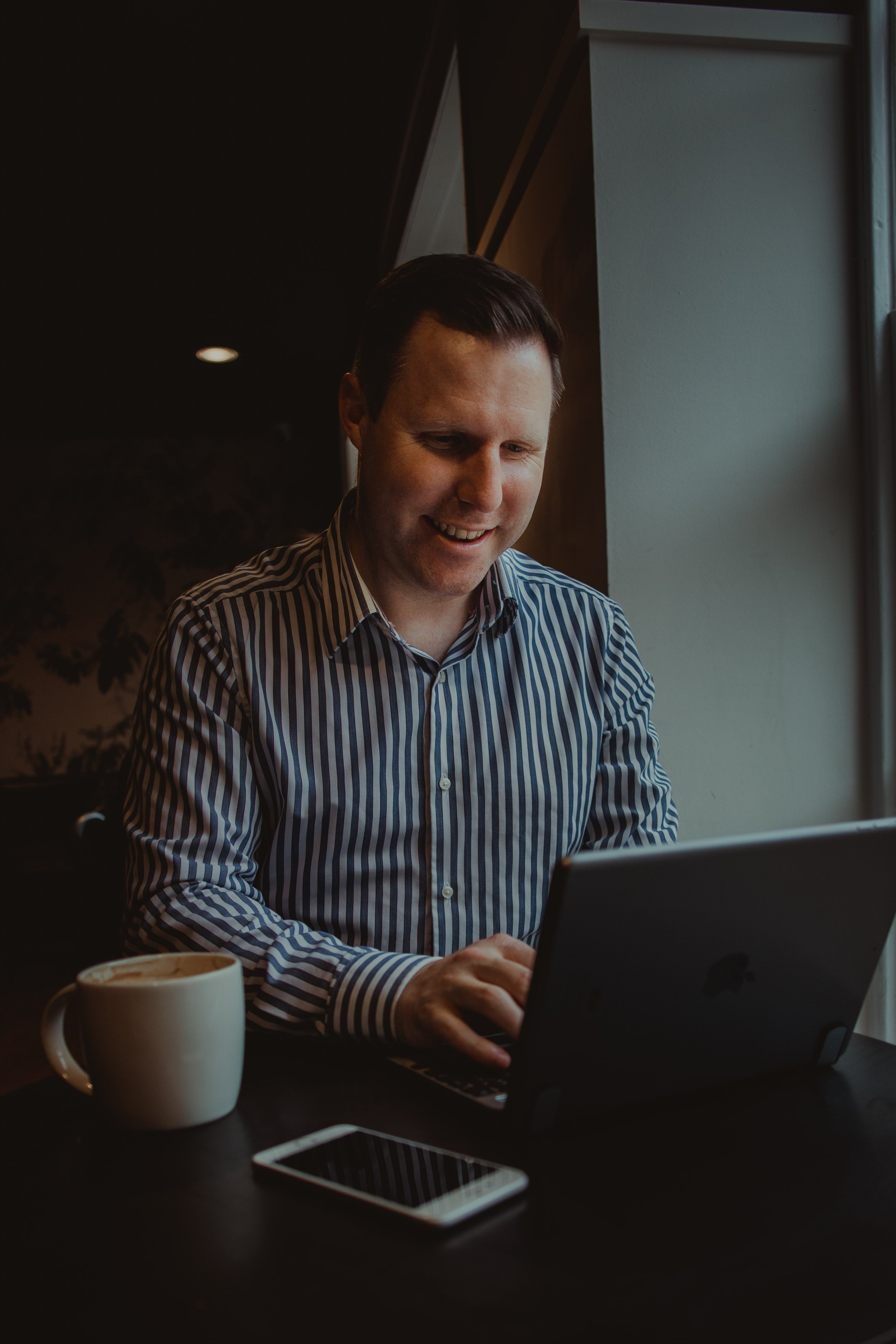 A man in a striped shirt smiling while working on a laptop at a dark wooden table near a window with natural light. There is a white mug with a beverage and a smartphone on the table.