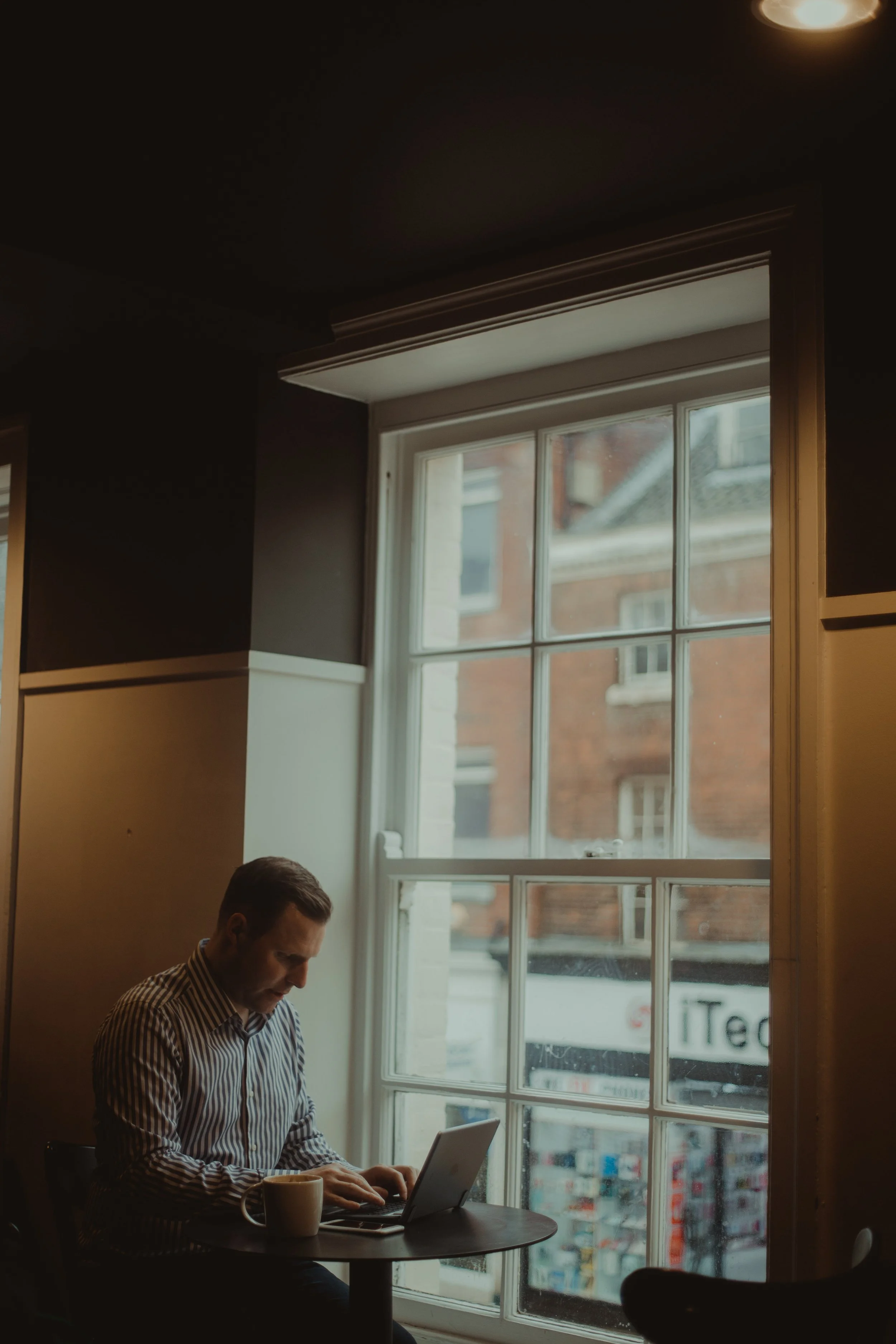 A man wearing a striped shirt working on a laptop at a small round table in a cozy cafe with large window, view of city buildings outside, and a coffee mug on the table.