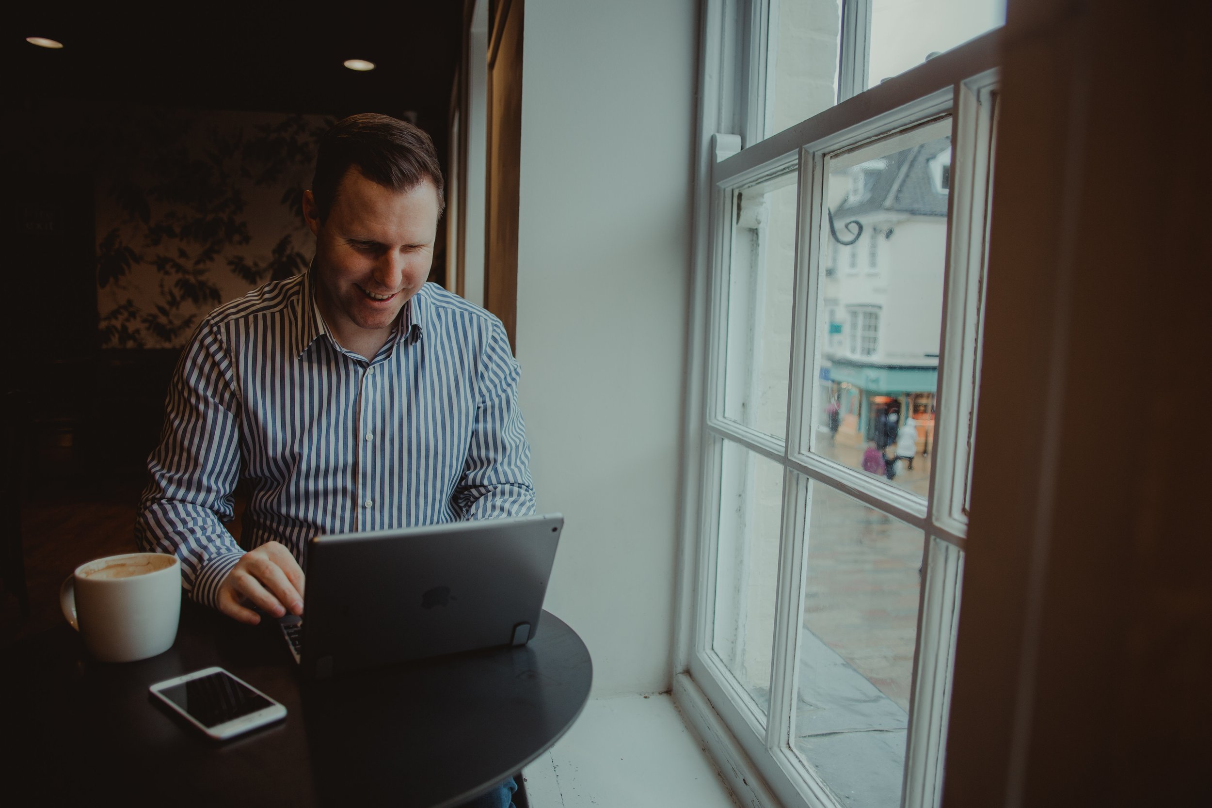 A man in a striped shirt smiling while working on a laptop at a table near a large window in a cozy cafe, with a cup of coffee and a smartphone on the table.