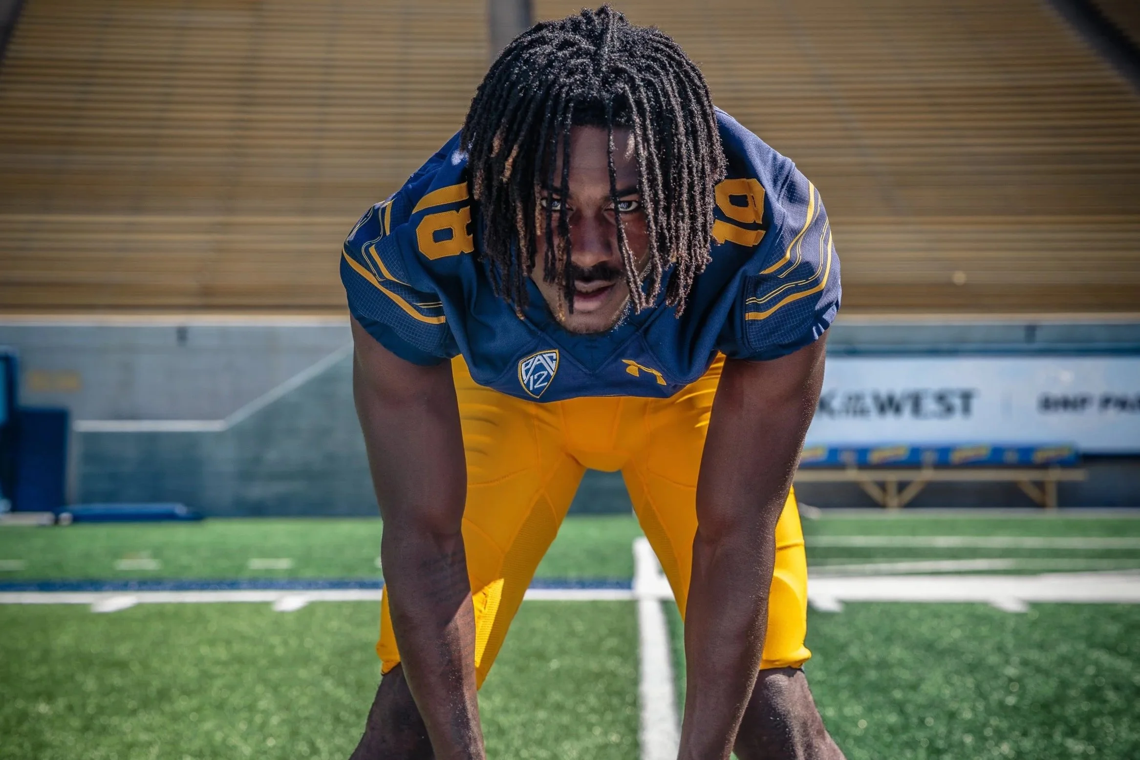 Football player in a blue jersey with yellow pants in a ready stance on the field.