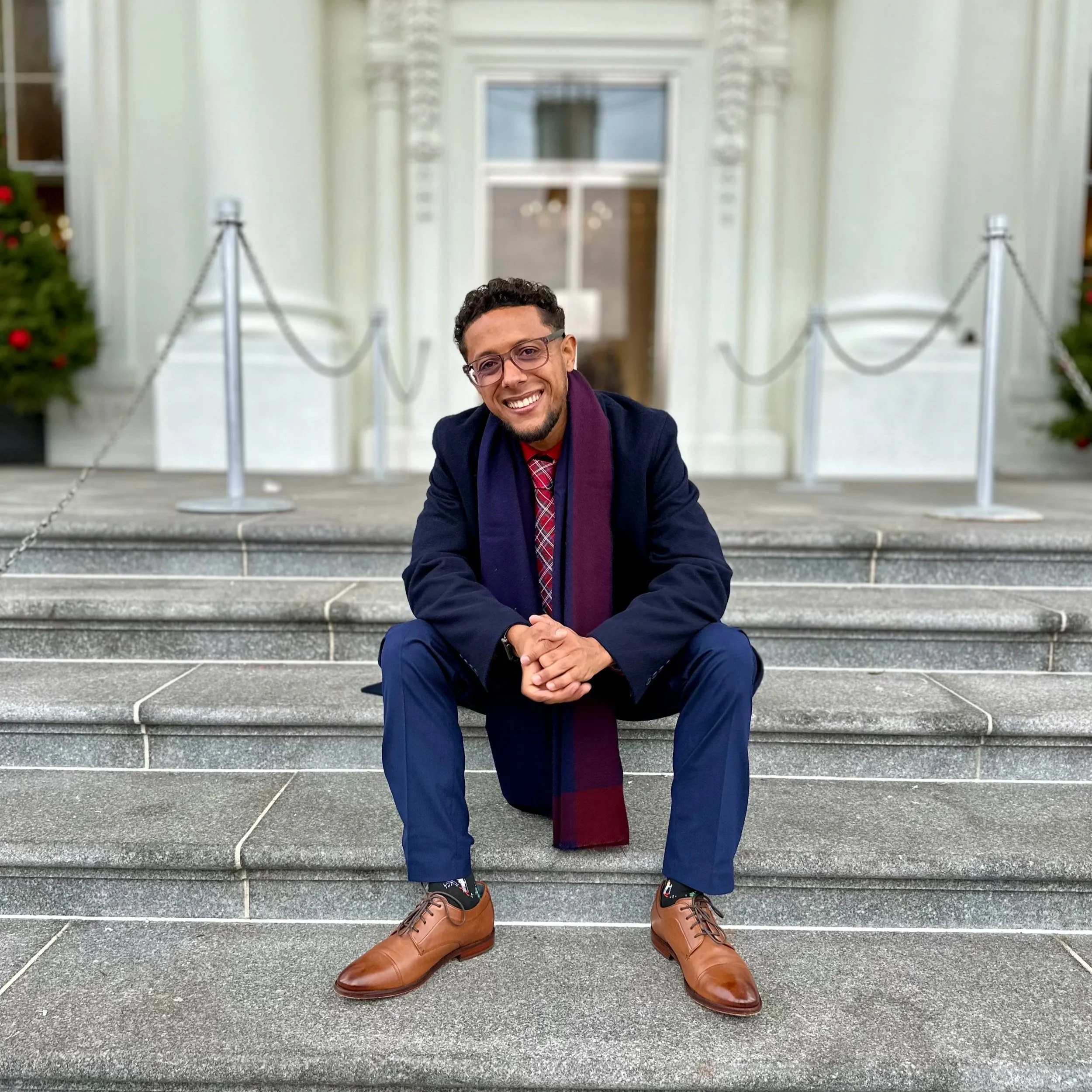 A man sitting on stairs smiling, wearing glasses, a navy blazer, a red tie, brown shoes, and a maroon scarf, with Christmas trees on either side of him and a white building with a door and window in the background.