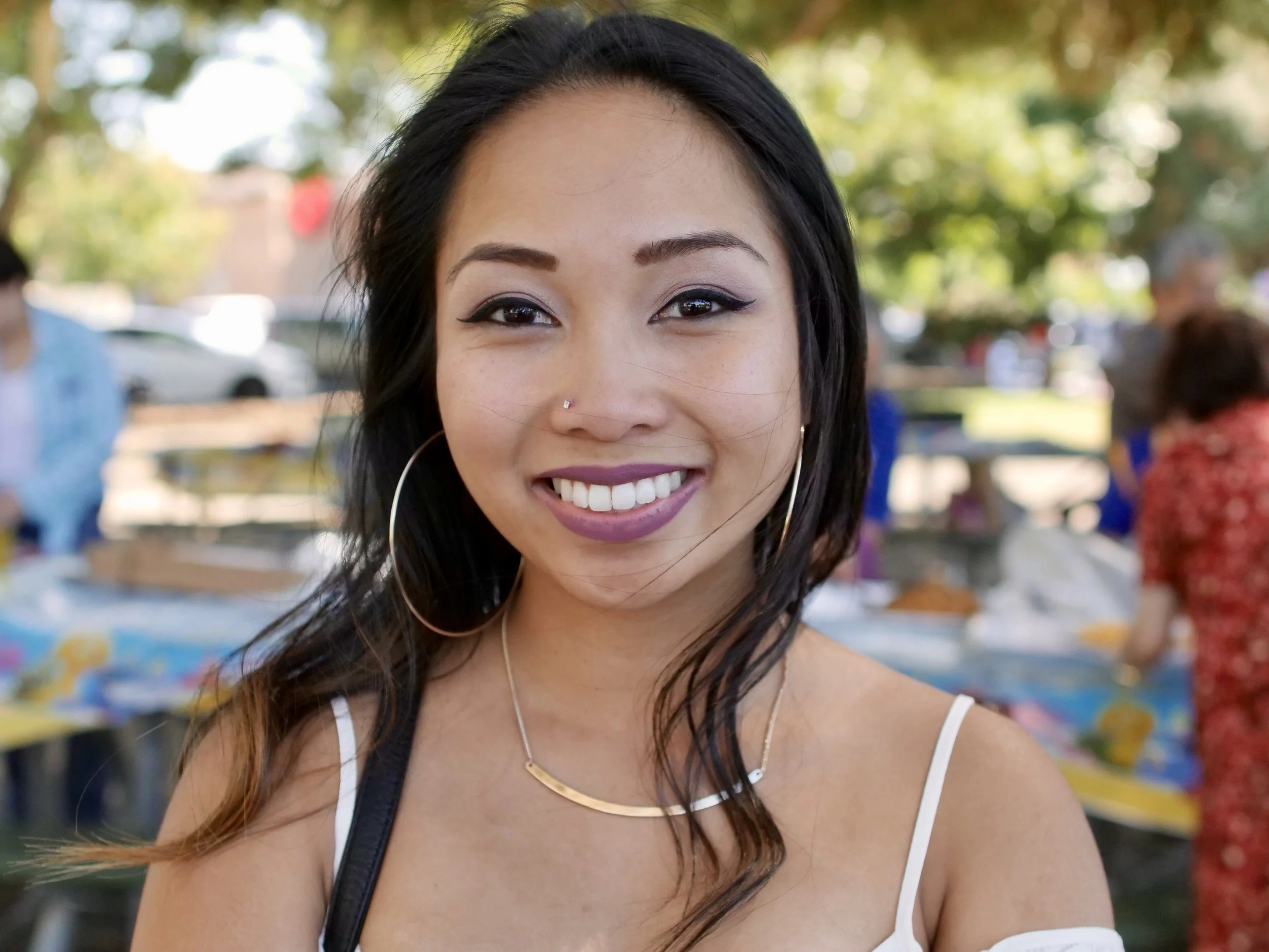 Close-up of a smiling woman with long dark hair, wearing hoop earrings and a thin gold necklace, outdoors on a sunny day with trees and blurred people in the background.