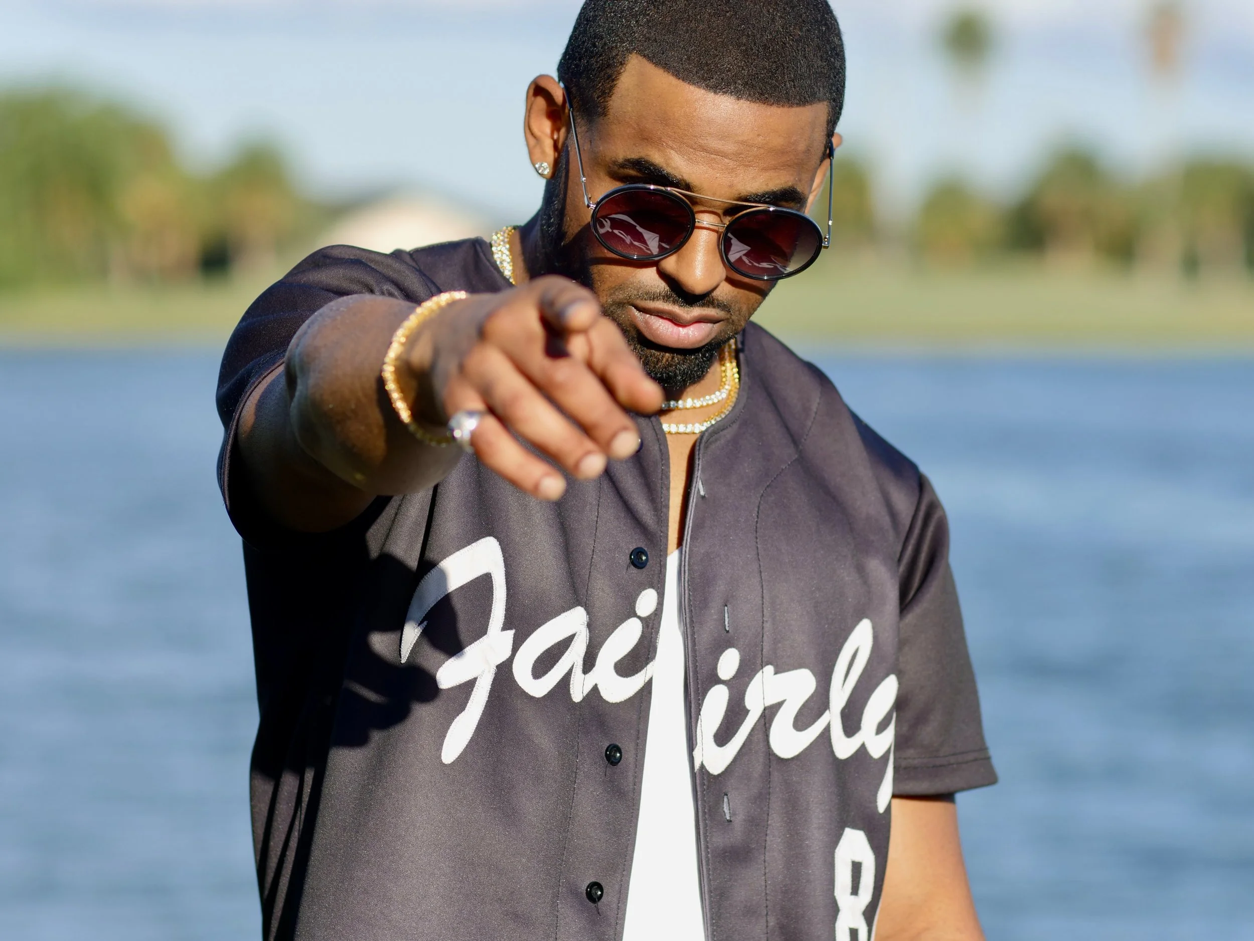 A man wearing sunglasses, gold jewelry, and a dark jacket with white cursive writing, pointing towards the camera in front of a body of water outdoors.