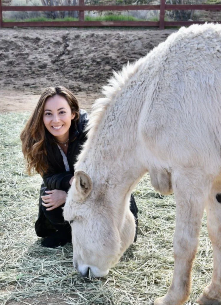A woman with long brown hair smiling and crouching beside a white horse in a paddock with hay and a wooden fence.