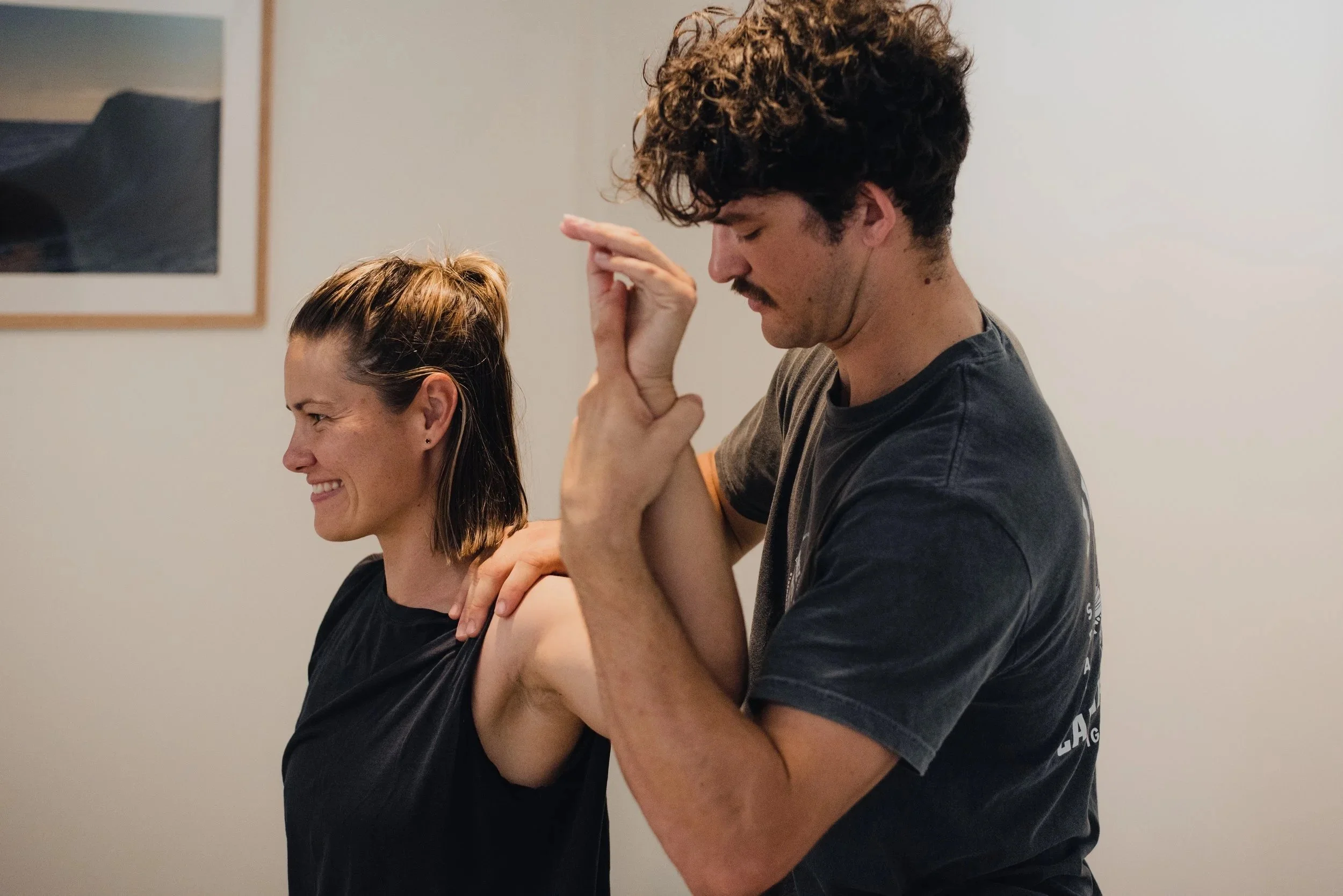 A man with curly hair is performing a massage on a woman with brown hair, who is smiling with her eyes closed. Both are wearing casual dark shirts, and the background is a plain light-colored wall with a framed picture.
