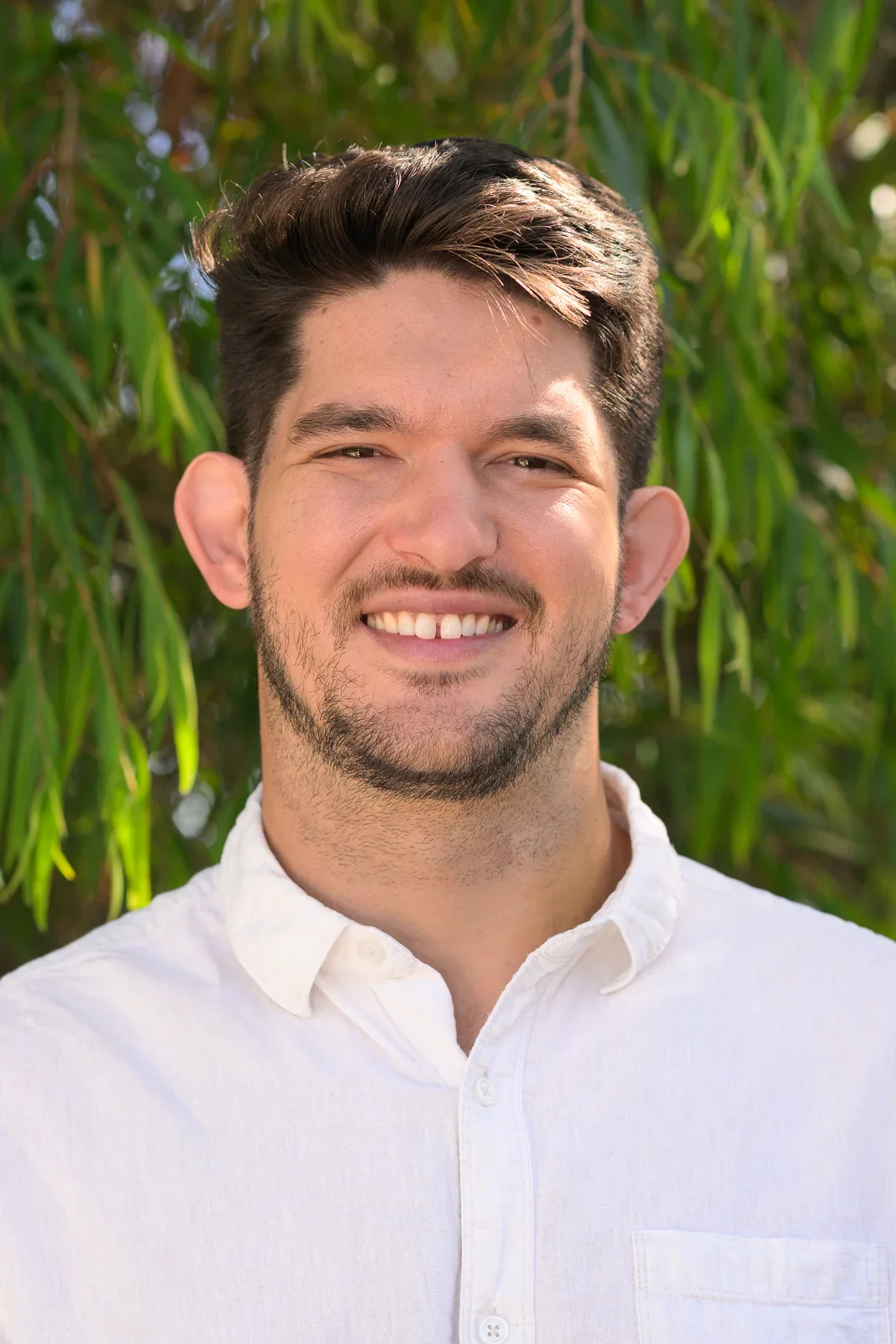 A man smiling outdoors with green foliage in the background, wearing a white button-up shirt.