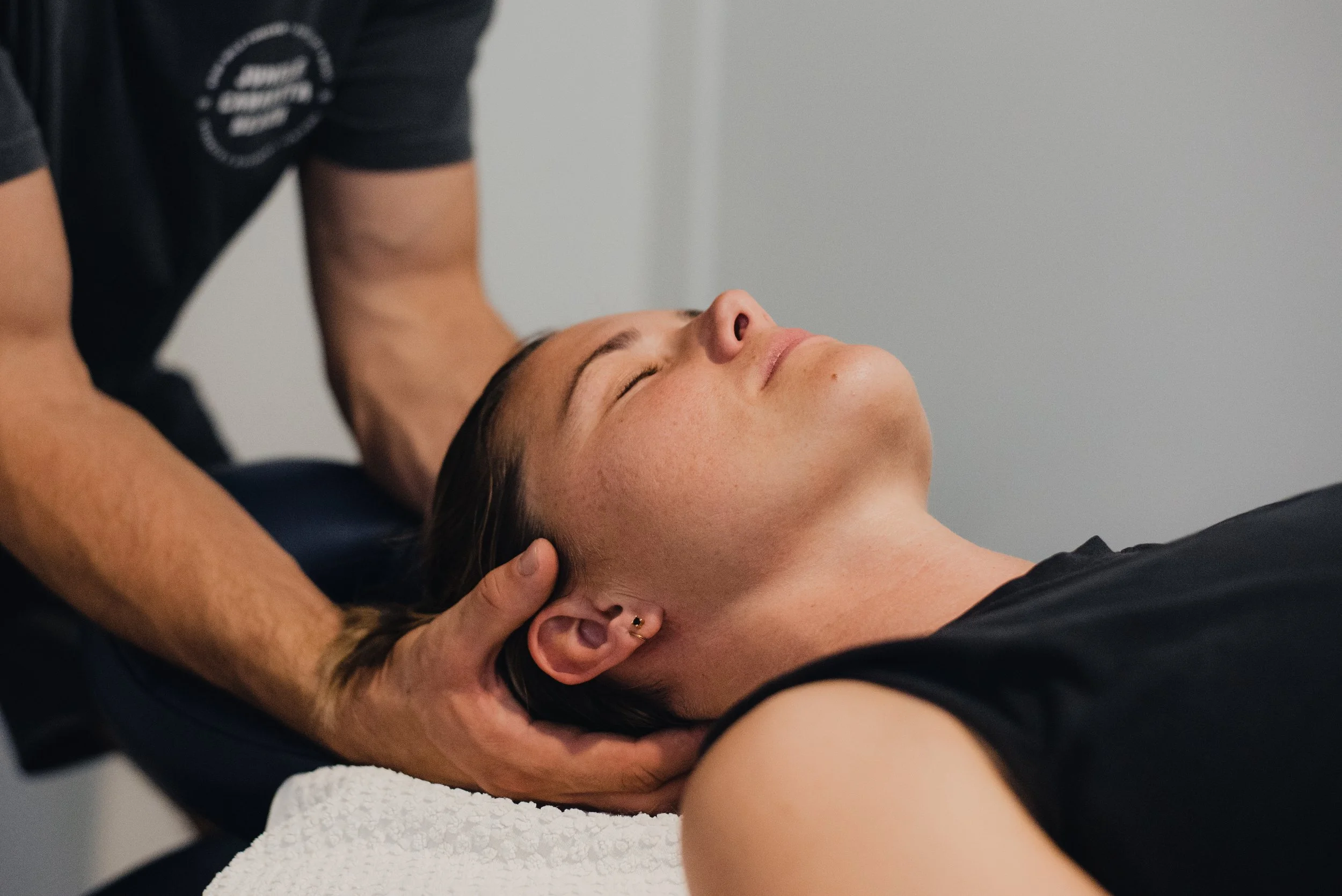 A woman receiving a massage while lying on her back with eyes closed, head tilted slightly to the side, and someone providing treatment with hands on her neck and shoulder area.