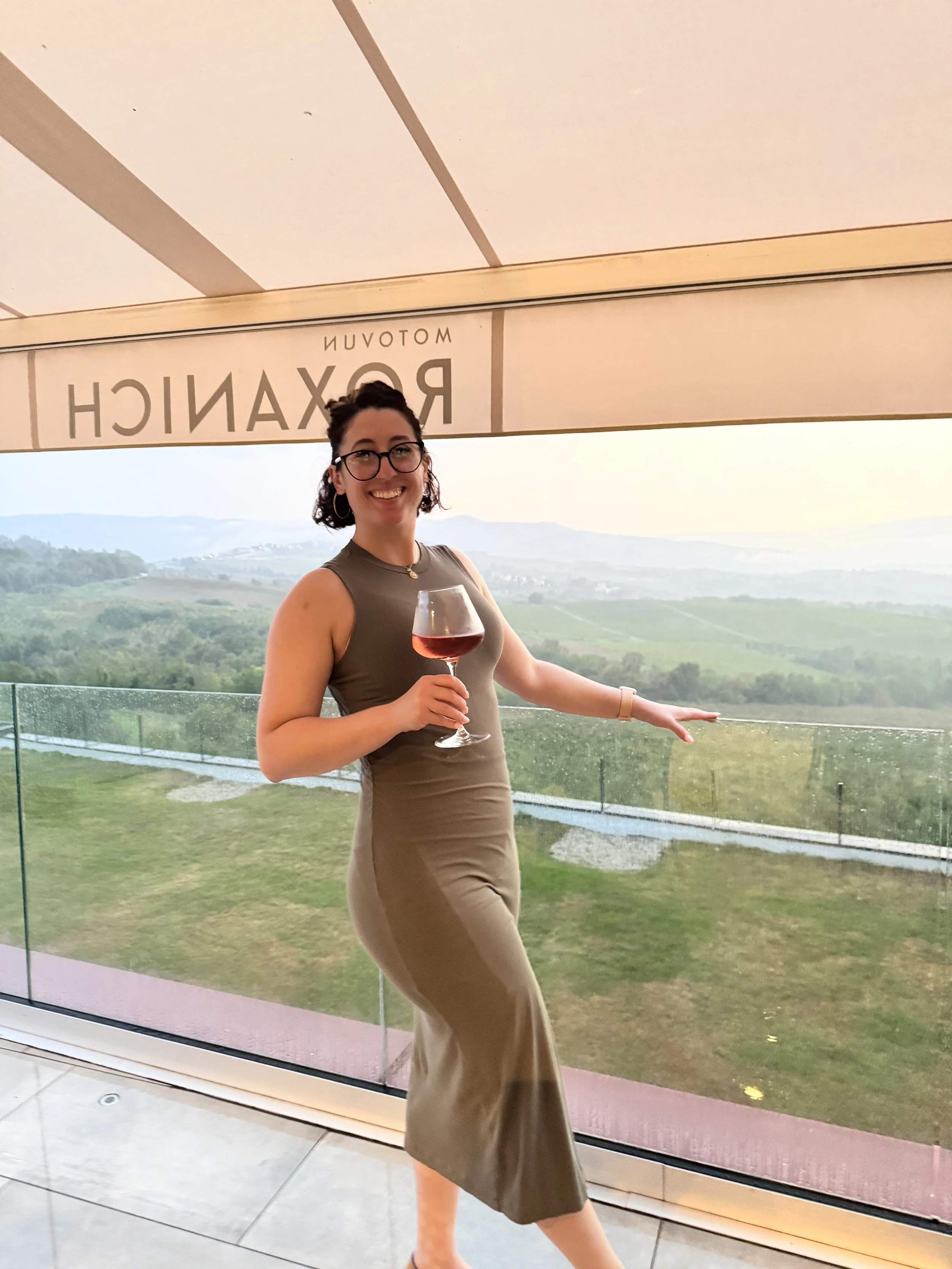 A woman with glasses and earrings smiling and holding a glass of red wine, standing in front of a large window with a scenic view of hills and countryside during sunset.