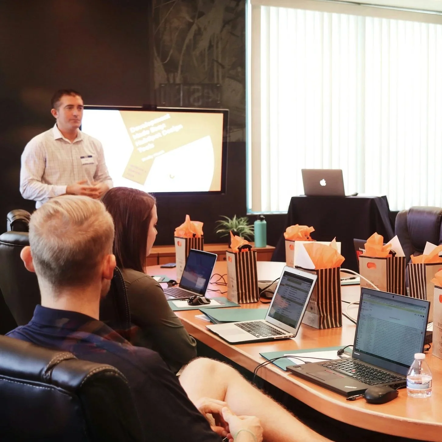 A presentation or meeting in a conference room with multiple people seated at a table with laptops. A man is standing near a large screen, giving a presentation. The table has gift bags with orange tissue paper on top. The room has large window blinds and a laptop on a small table.