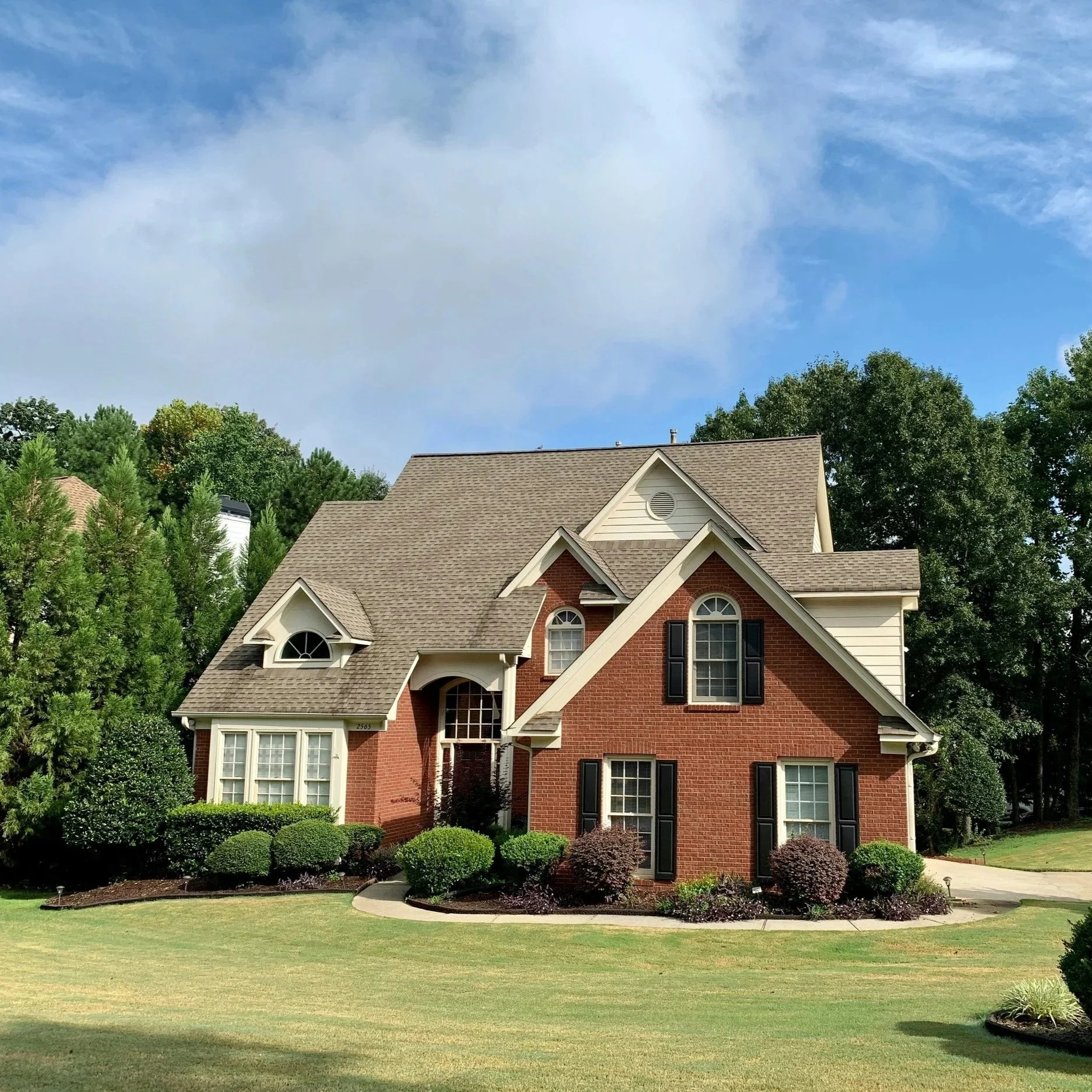 A large brick house with multiple gabled roofs, large windows, and black shutters surrounded by a well-manicured lawn and bushes, with a partly cloudy blue sky overhead.