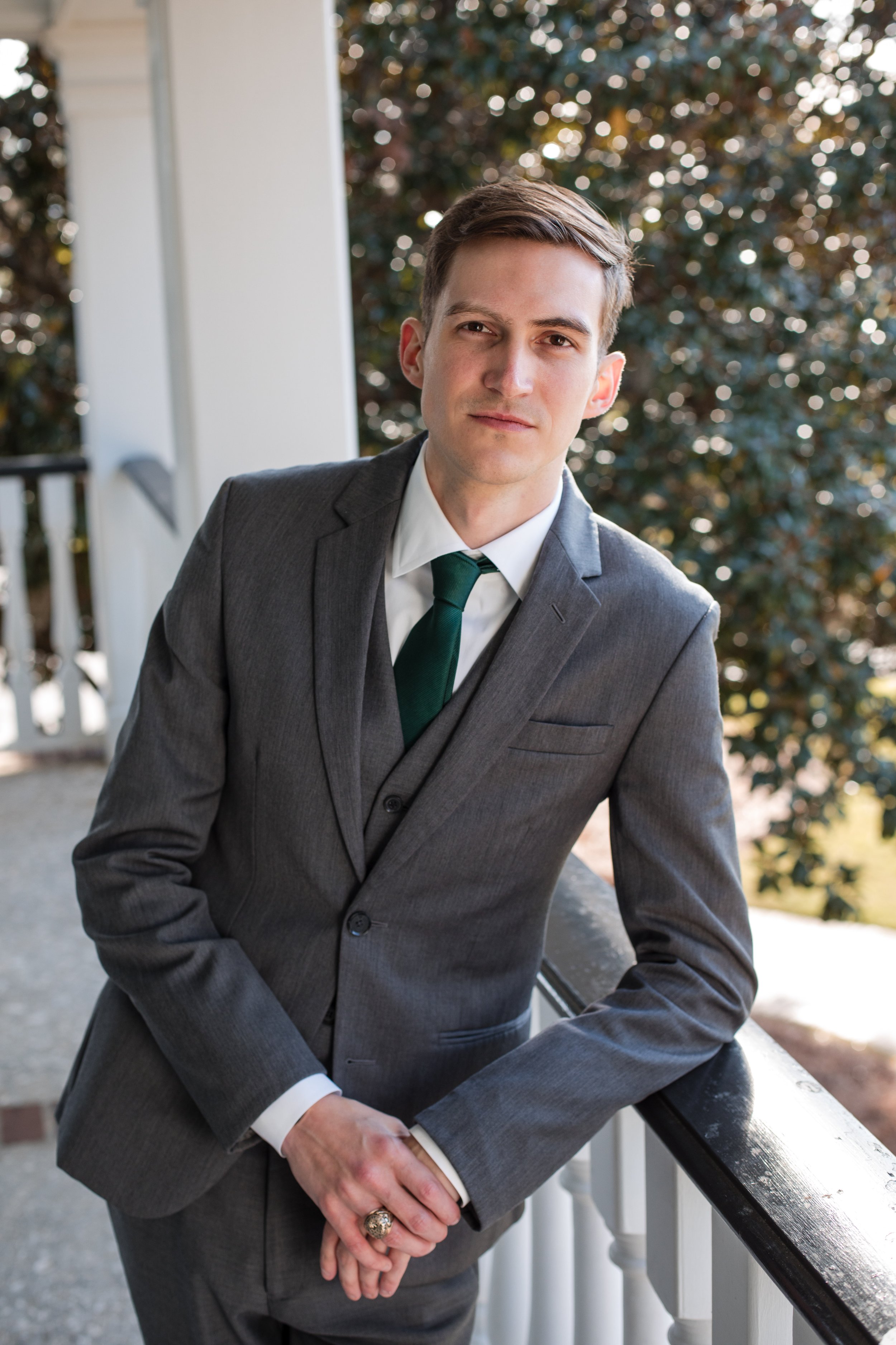 A young man in a gray suit with a green tie, standing outdoors on a porch railing, with trees and sunlight in the background.