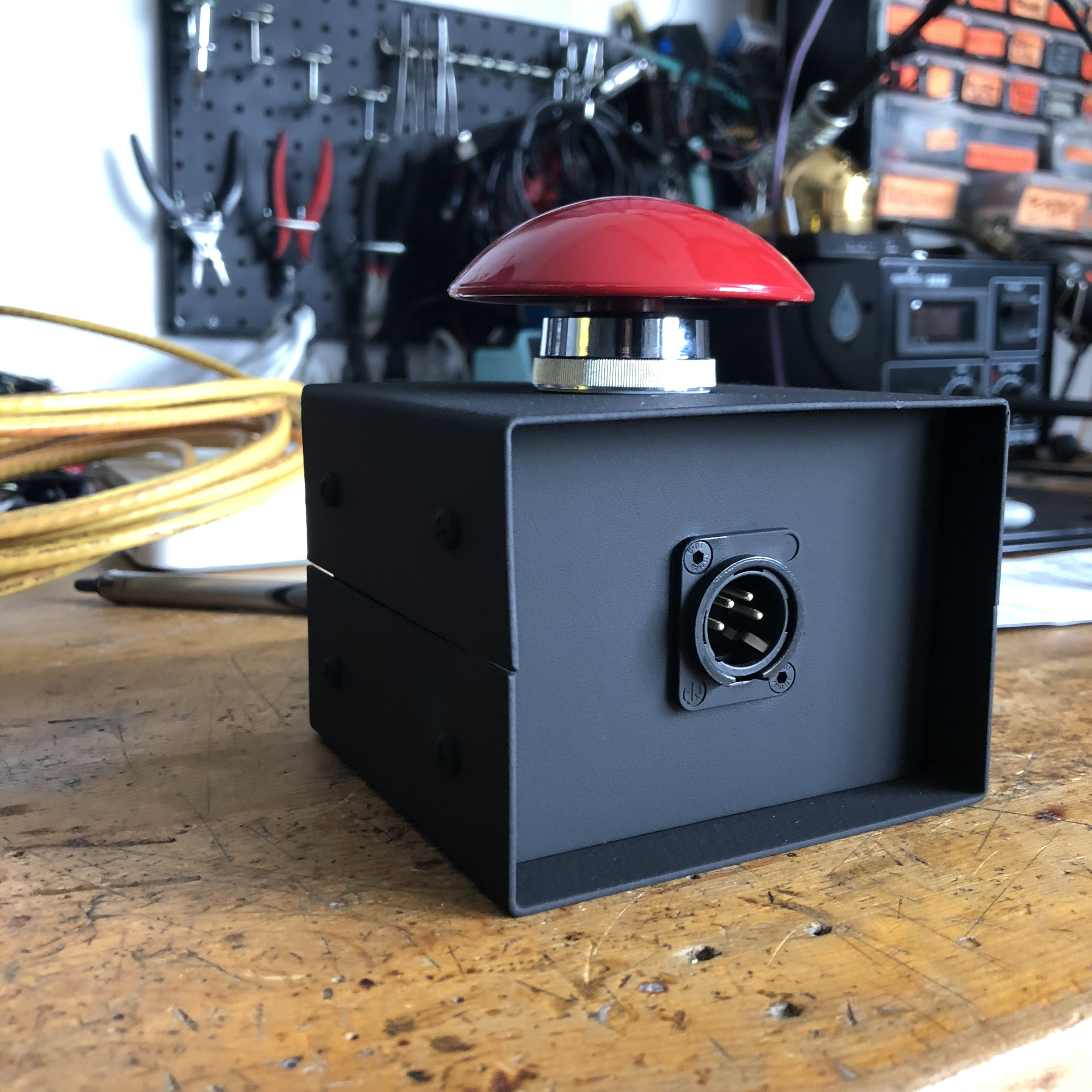 A close-up of a red button with a metallic ring, mounted on a black box, with various tools and supplies in the background, on a wooden surface.