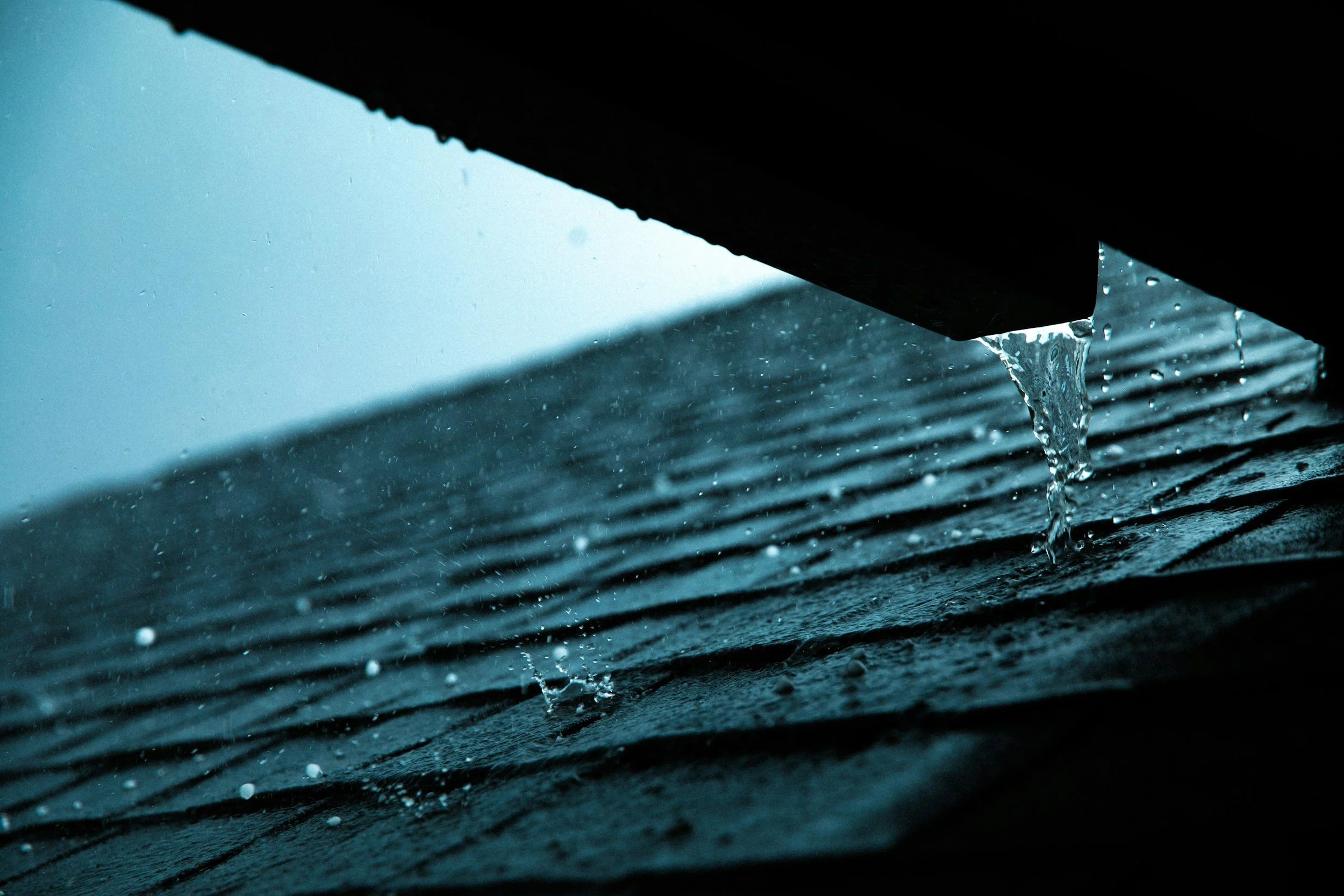 Close-up of water dripping from a dark surface, with the background showing rain on a window.
