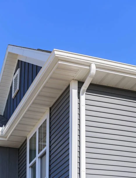 Close-up of the corner of a house with gray siding, white trim, and a white gutter system under a clear blue sky.