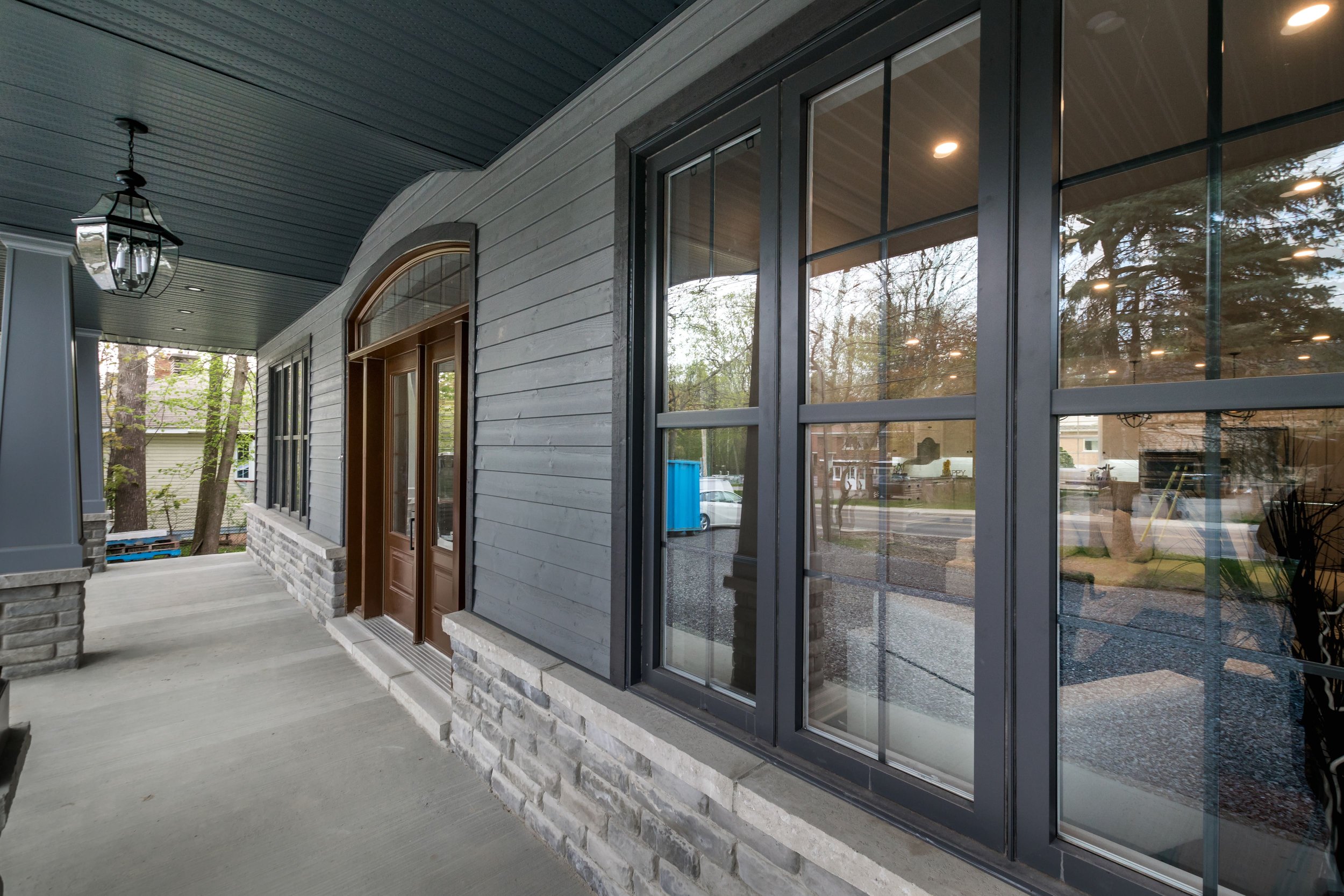 Front porch of a house with large windows, gray siding, brick accents, and porch lights, in a suburban neighborhood.