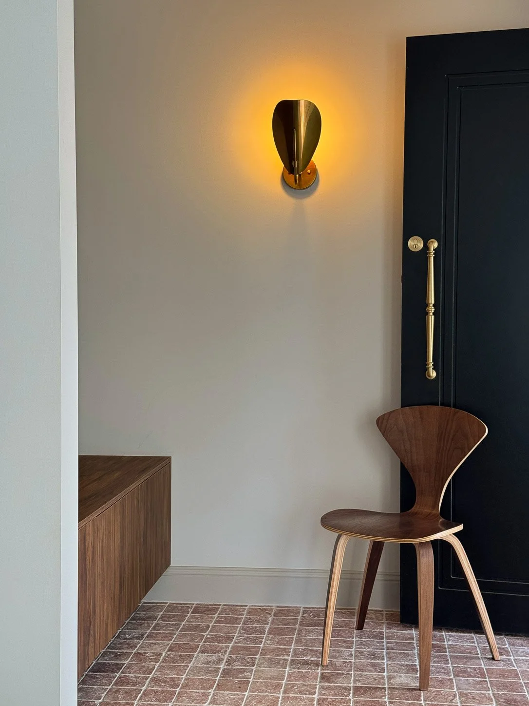 Warm entry at Falls House with walnut chair, dark timber door and brass wall light.