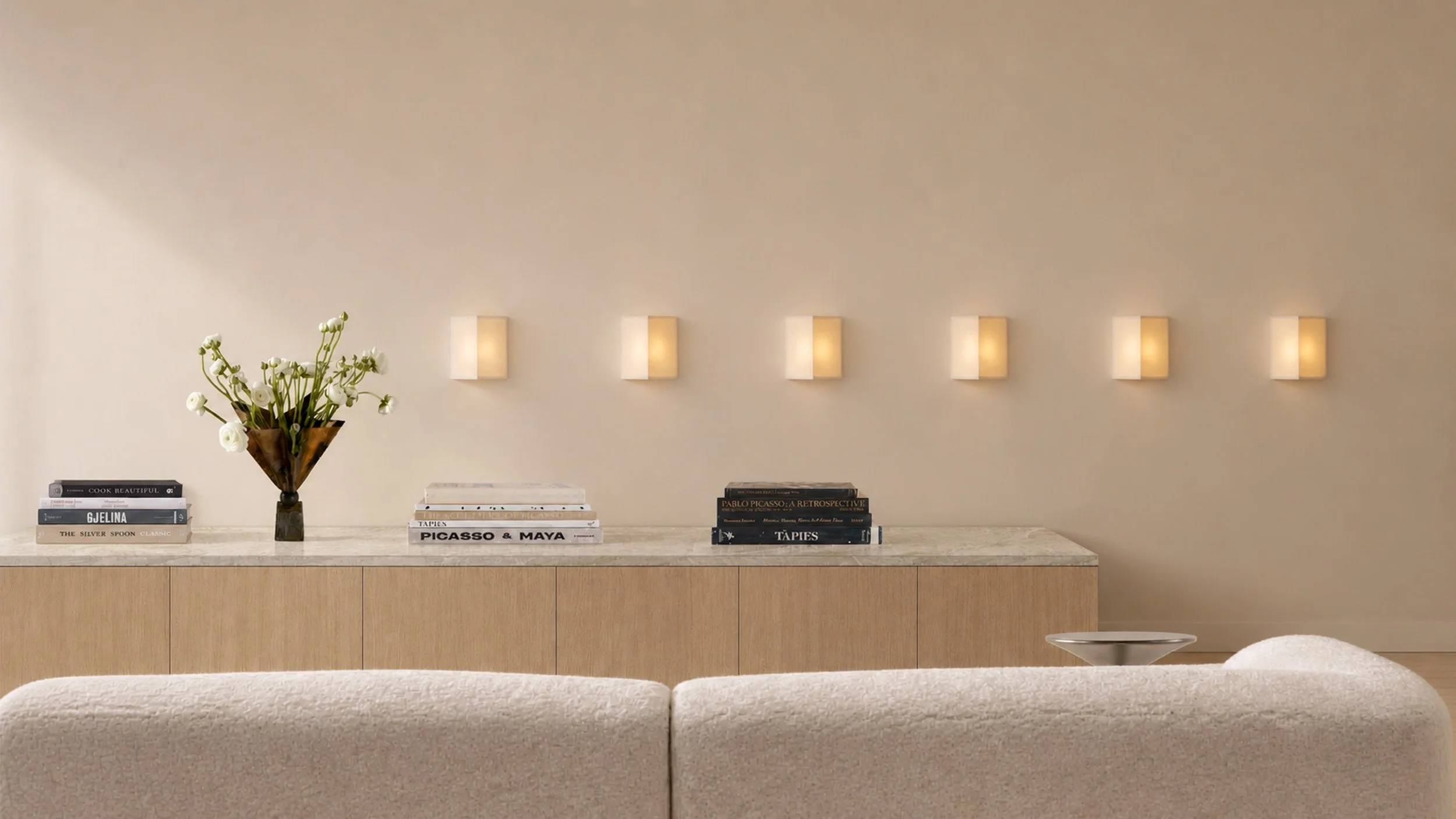 A row of six linen sconces above an oak credenza with art books and white ranunculus