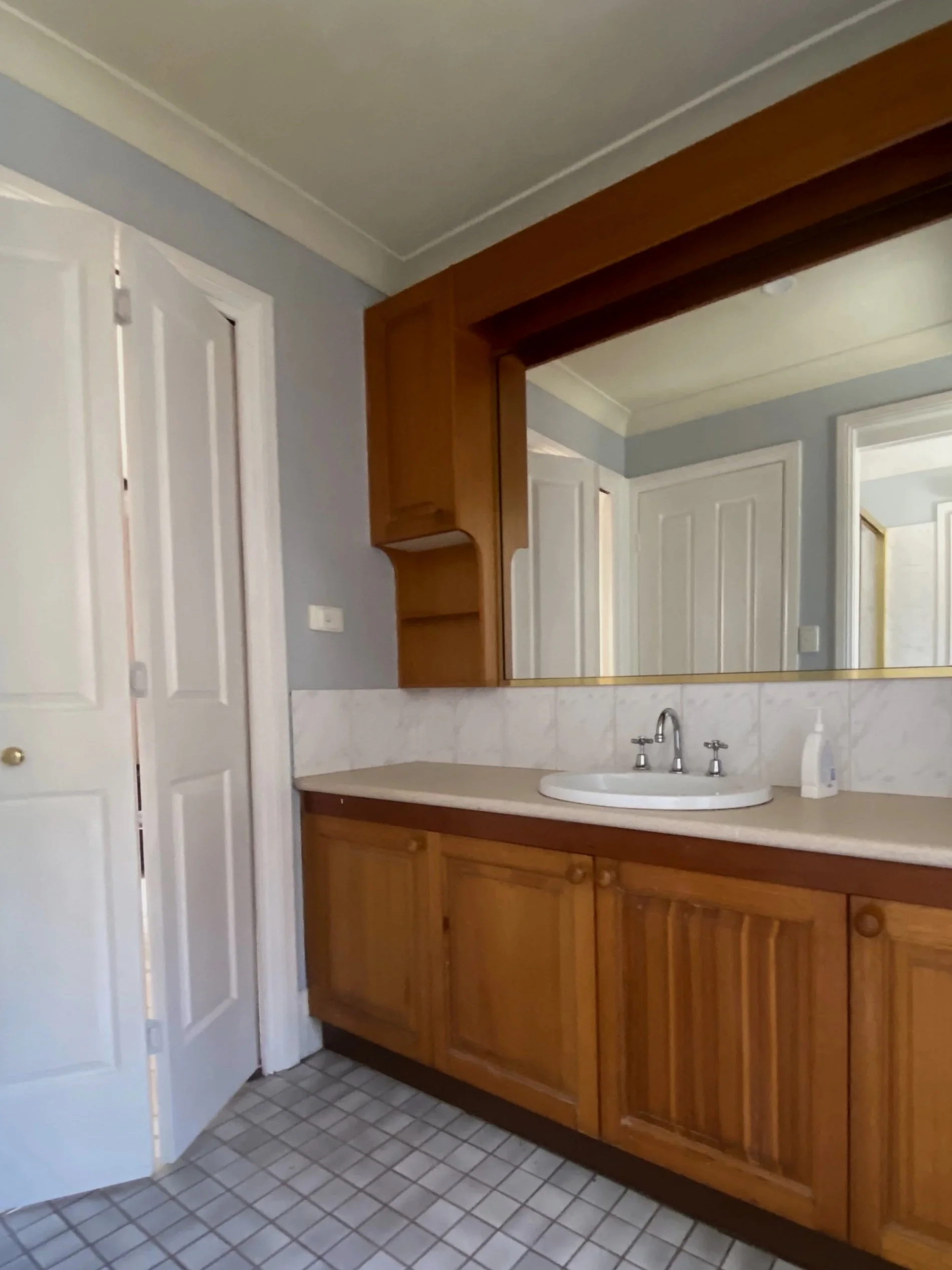 Old bathroom vanity with wooden cabinets, a white sink, faucet, and mirror, with tiled floor and baby blue walls.