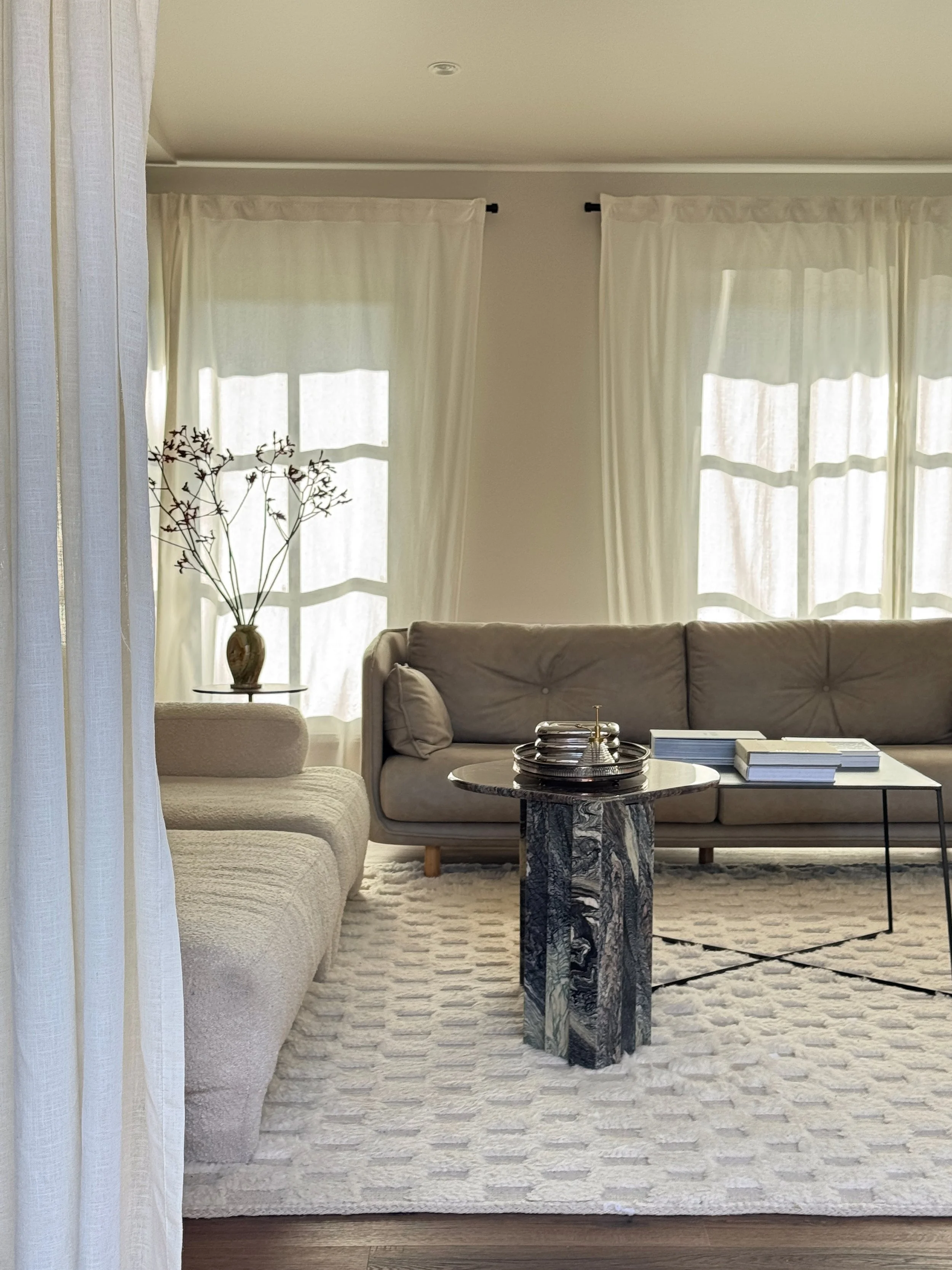 Living room with ivory and beige sofas, a set of nestled coffee tables of marble and steel, a decorative vase with branches, and large windows with sheer curtains, illuminated by natural light.