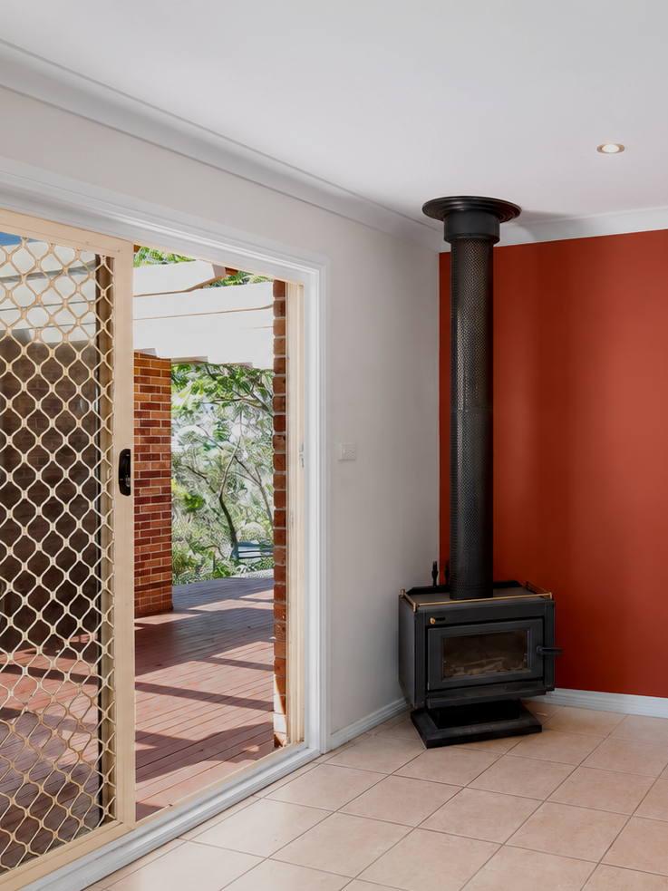 A cozy interior room with tiled floor, white walls, a red accent wall, a wood-burning stove, and a sliding glass door leading to an outdoor patio with brick and wooden surroundings.
