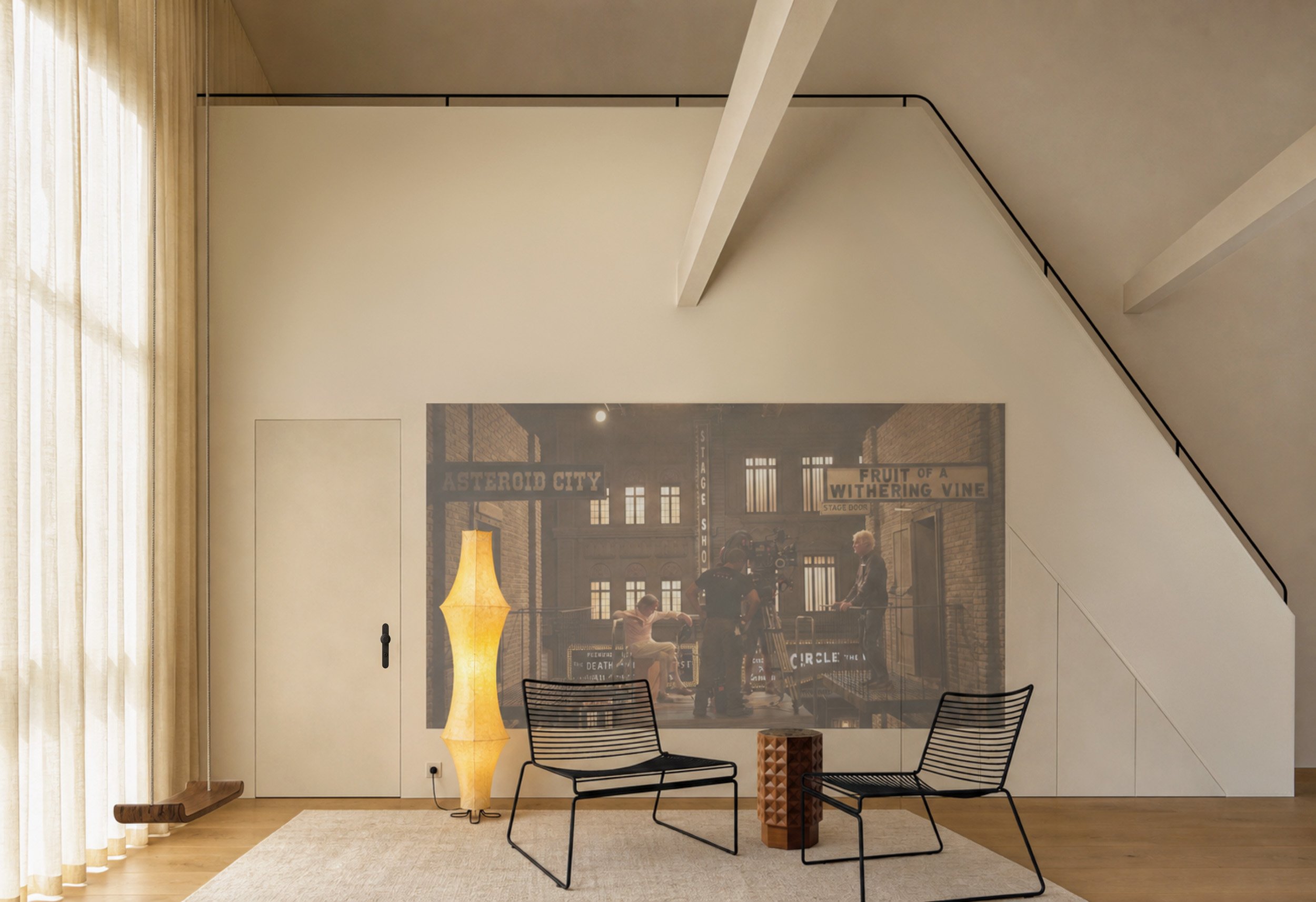 Modern loft interior with black chairs, a tall glowing paper floor lamp, a small wooden side table, and a projected movie scene of a city street scene on the wall.