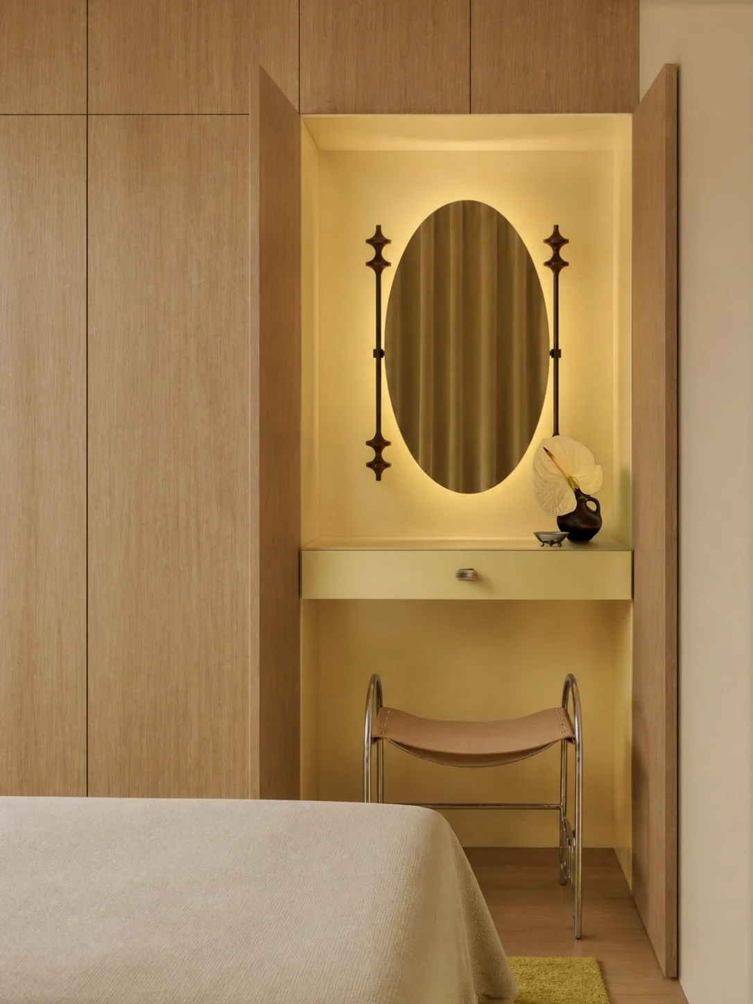 Dressing nook with a backlit oval mirror, bronze sconces and a chrome and leather stool