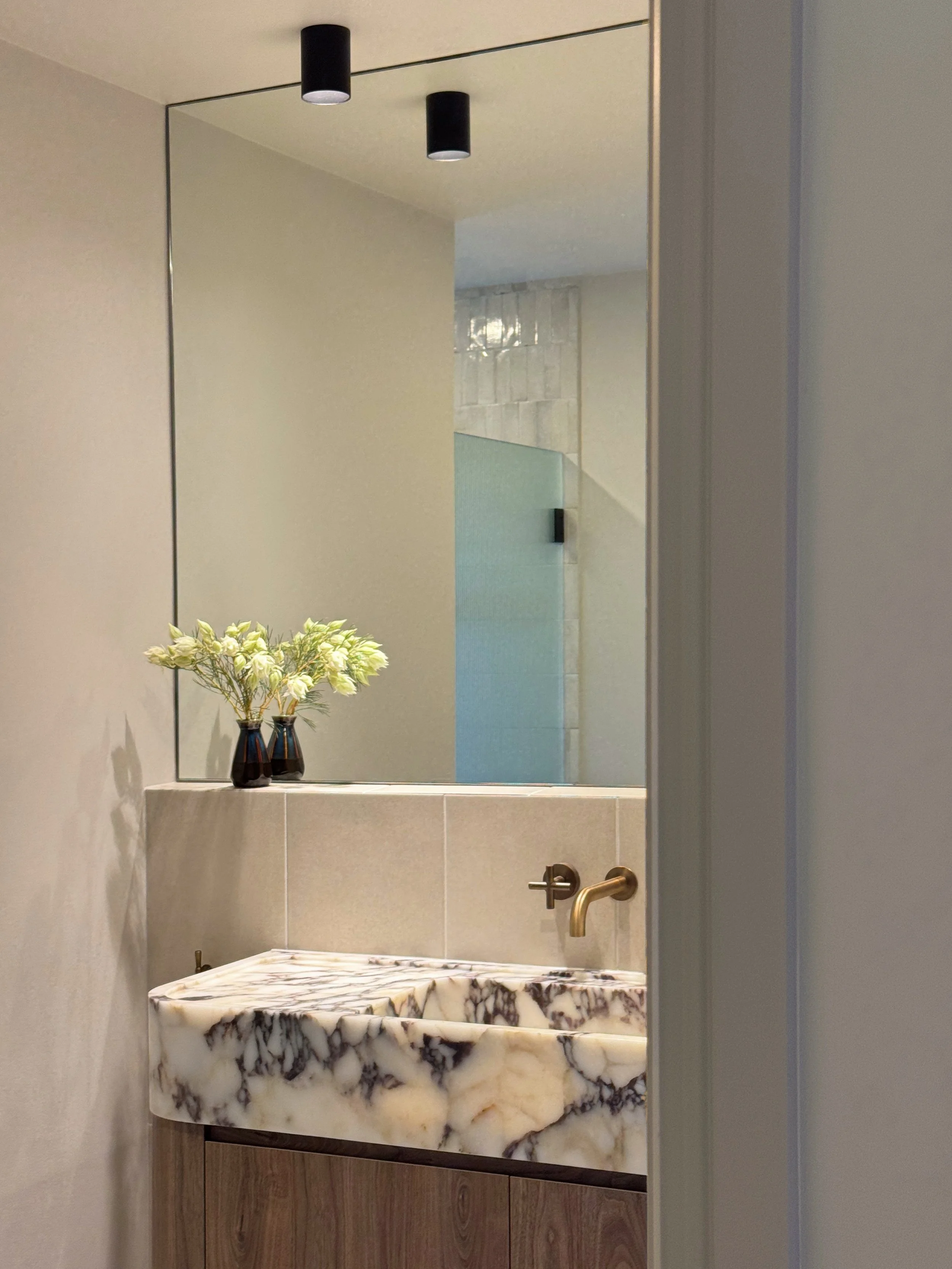 A threshold view of the renovated bathroom with a calacatta viola marble sink, a small ceramic vases with white flowers, a mirrored wall and a wall-mounted brass faucet.