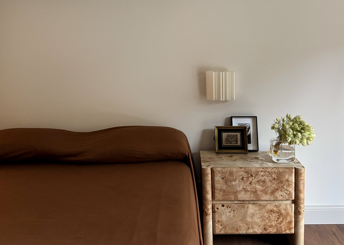 Bedside detail — burl wood table, fluted wall light and framed pictures against tobacco bedding