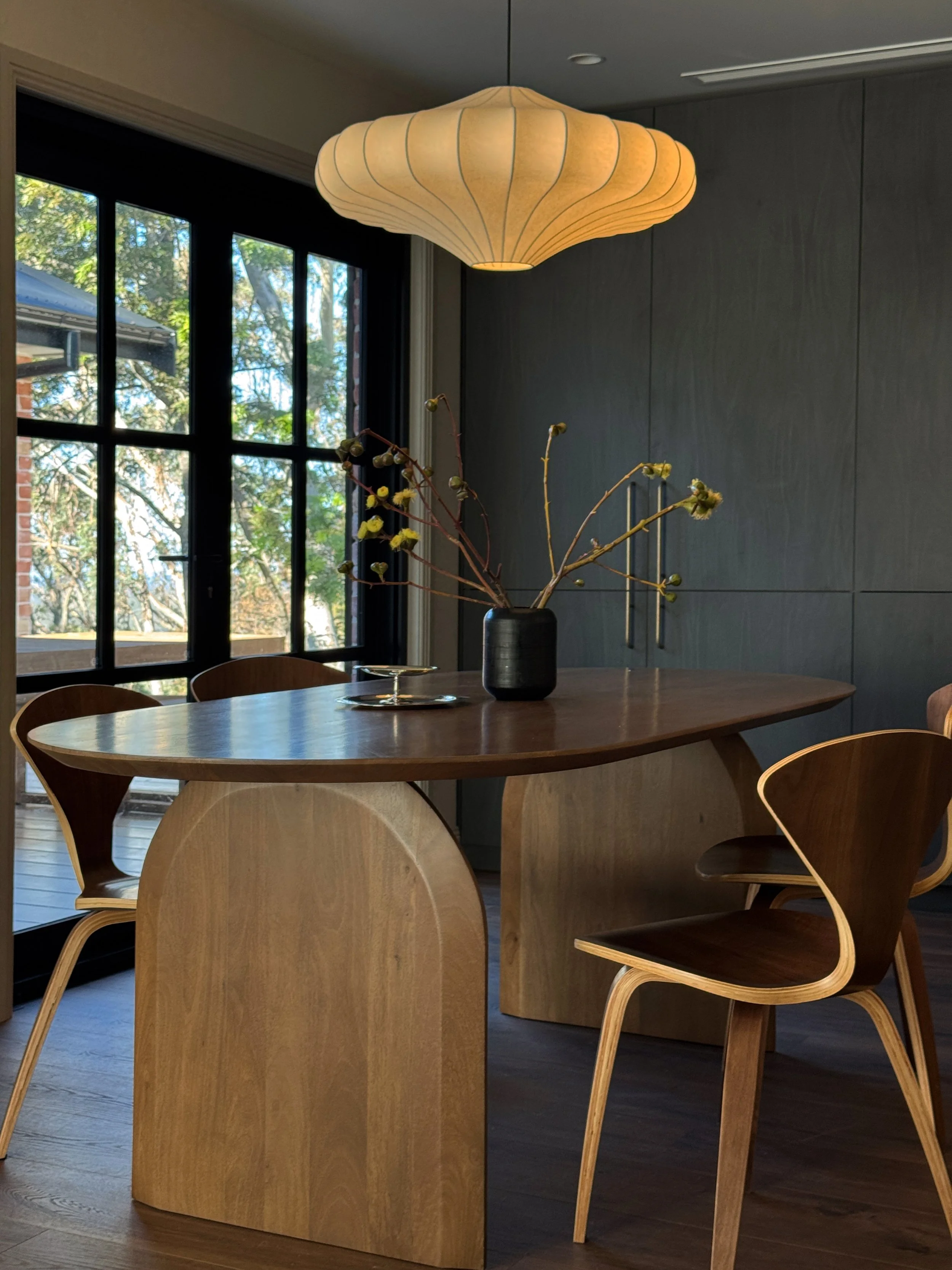 A round wooden dining table with four wooden chairs, a black vase with branches and buds, a small silver tray, and a large beige paper lantern hanging from the ceiling by a black cord inside a modern room with dark wooden floors and large black-framed windows.