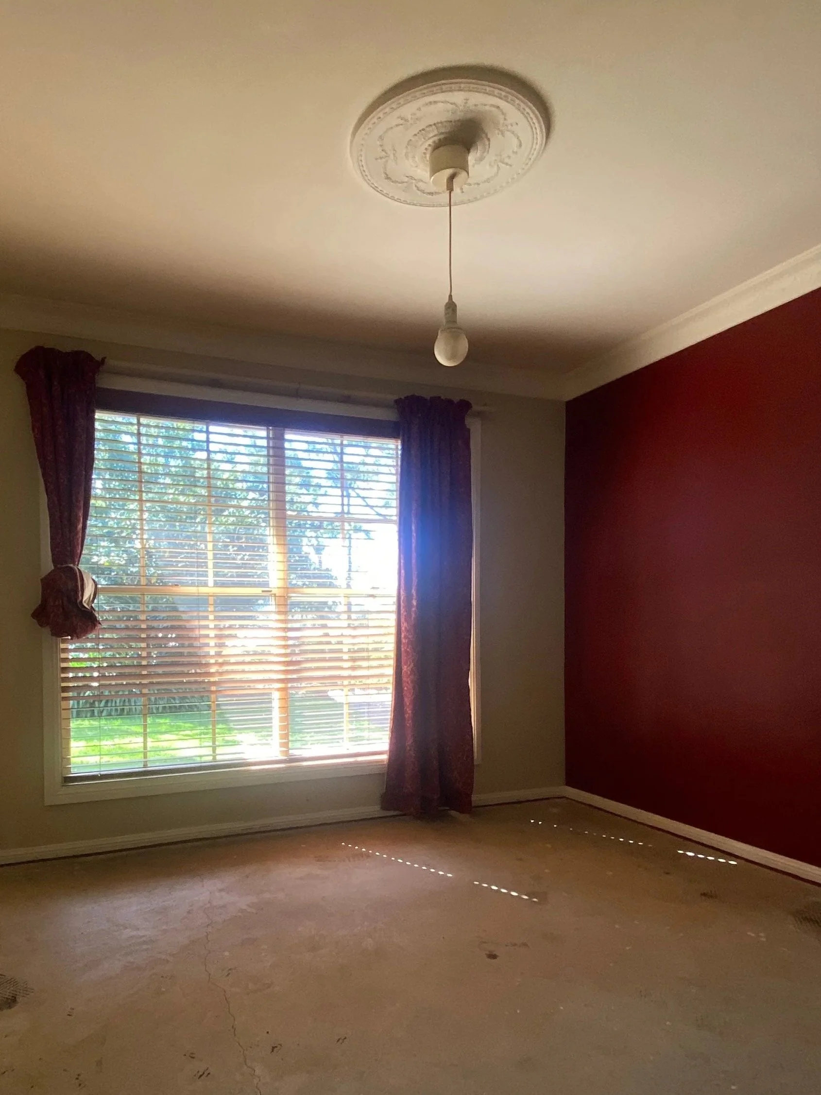 Old living room with red accent wall, and large window with closed blinds and maroon curtains.