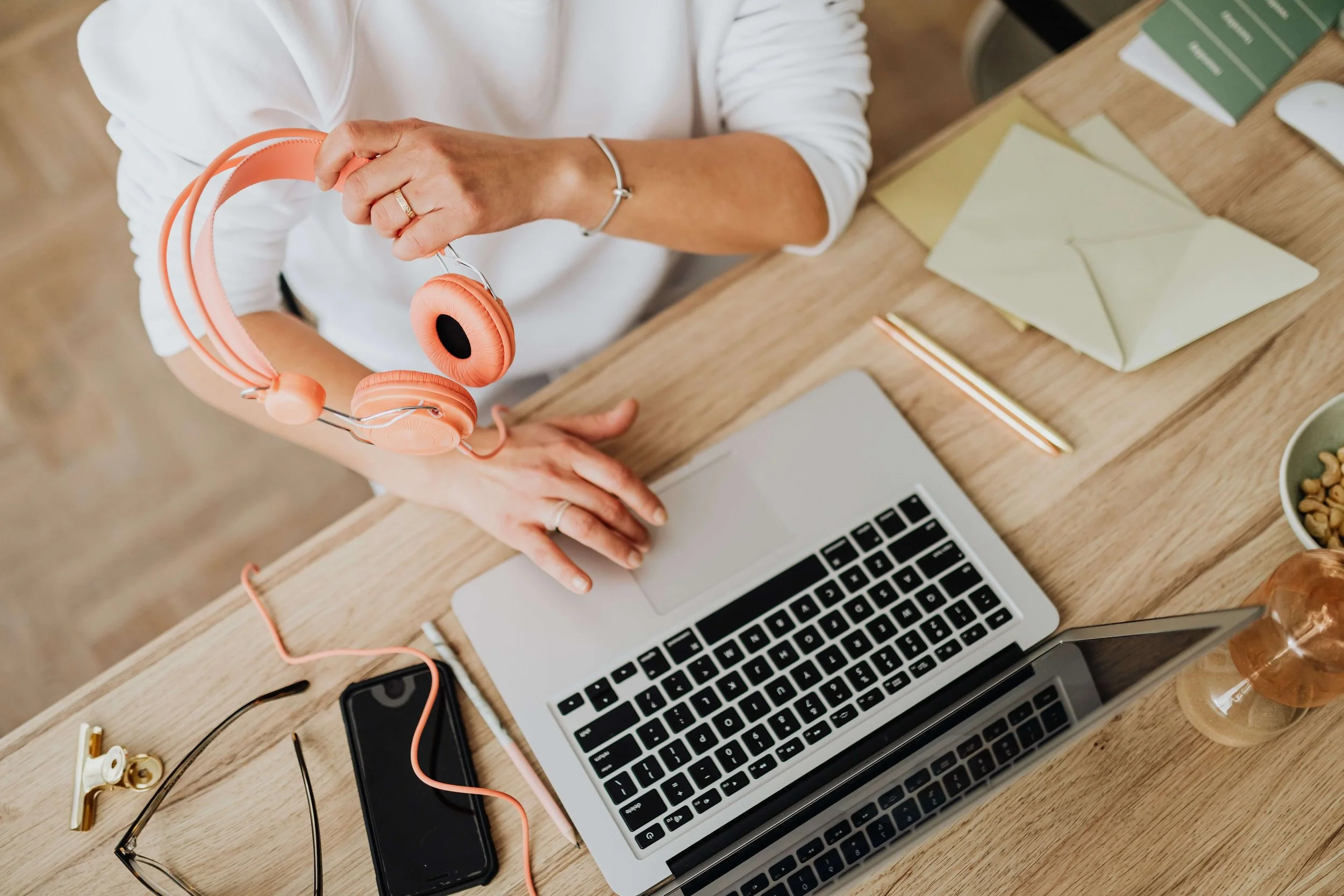 A person holding pink headphones over a laptop on a wooden desk surrounded by items including a smartphone, glasses, a pen, and a bowl of snacks.