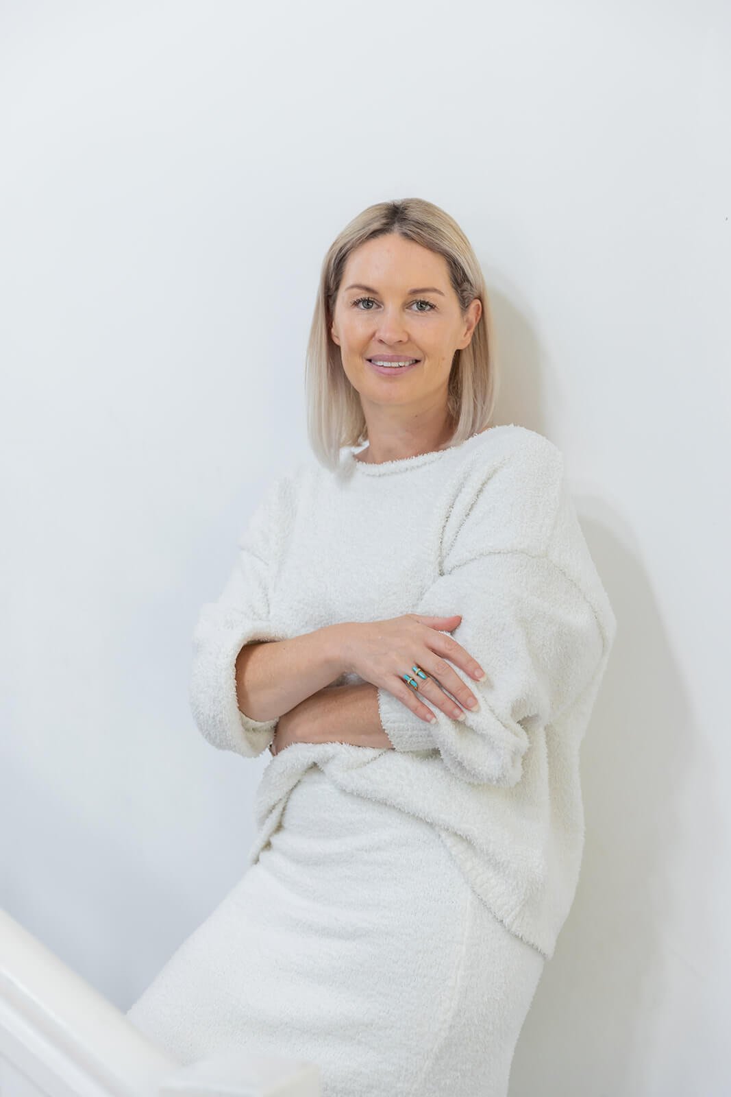 A woman with blonde hair wearing a white sweater and skirt, standing against a plain white wall, smiling and posing with arms crossed.