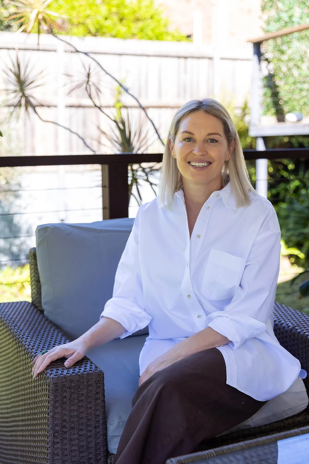 A woman with blonde hair wearing a white button-up shirt sitting outdoors on a wicker couch with a gray cushion and a wooden railing behind her, smiling at the camera.