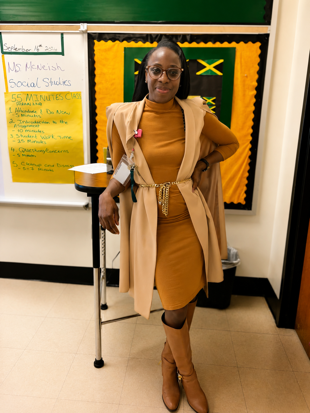 A woman standing next to a classroom chalkboard and a yellow schedule poster, dressed in tan clothing including a dress, trench coat, and knee-high boots, with glasses, black hair styled in a ponytail, and a confident expression.