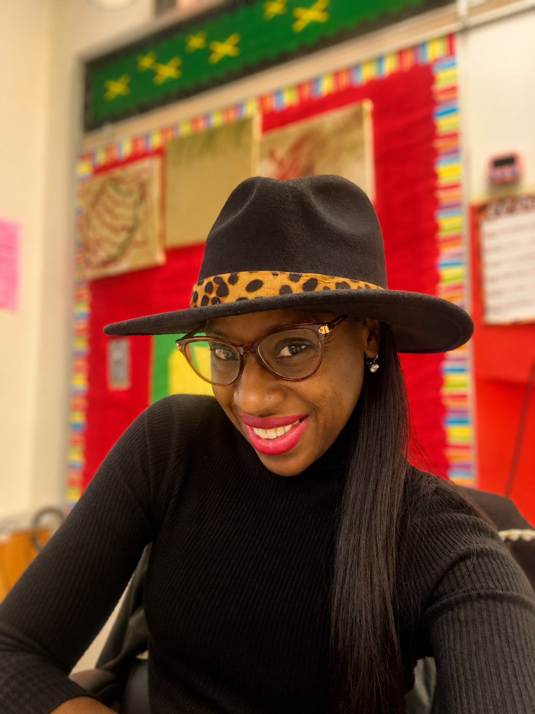 A woman with long dark hair, wearing glasses, a black hat with a leopard print band, and a black top, smiling at the camera inside a classroom with colorful posters and decorations on the wall.