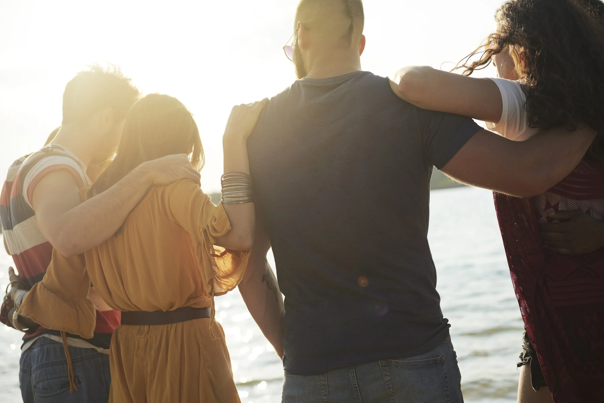 A group of friends stand close together with arms around each other, facing the water during sunset on a beach.