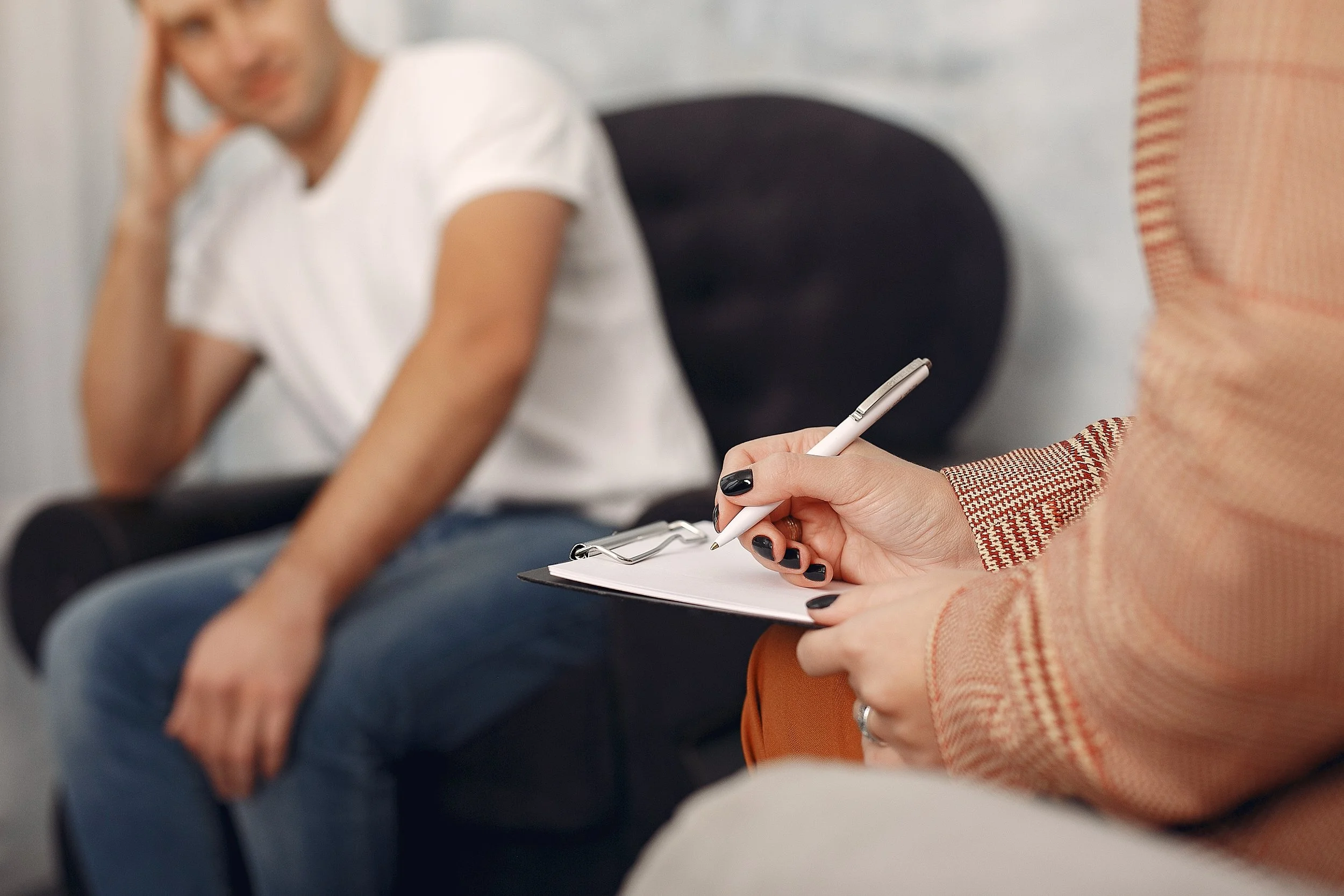 A woman in a plaid blazer taking notes on a clipboard during a meeting, with a man in a white shirt listening in the background.