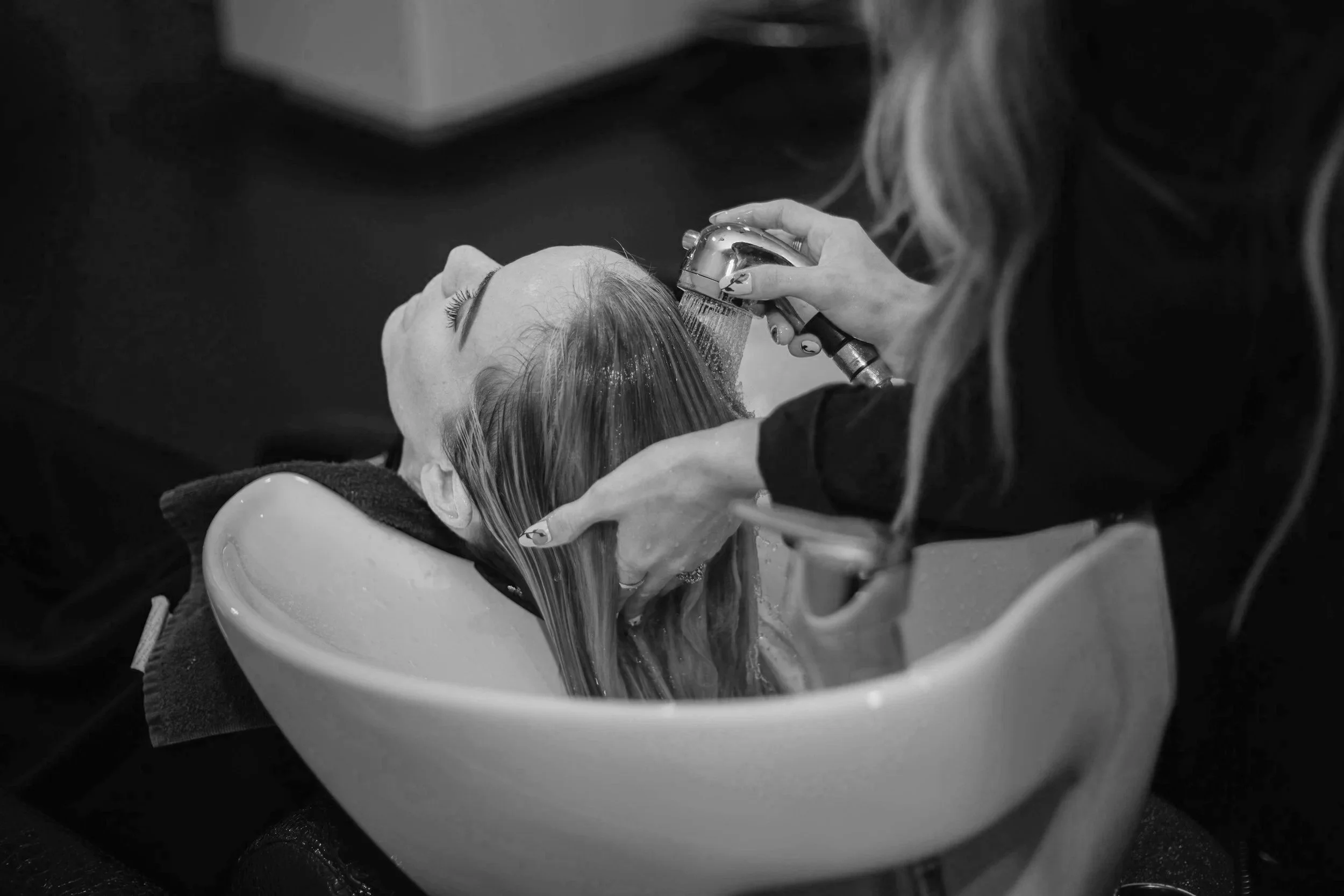 A woman is lying back in a salon chair while receiving a hair wash. A stylist is rinsing her hair with a handheld showerhead over a sink. NHR Studios | Nashville Hair Replacement | J. Bangs Salon