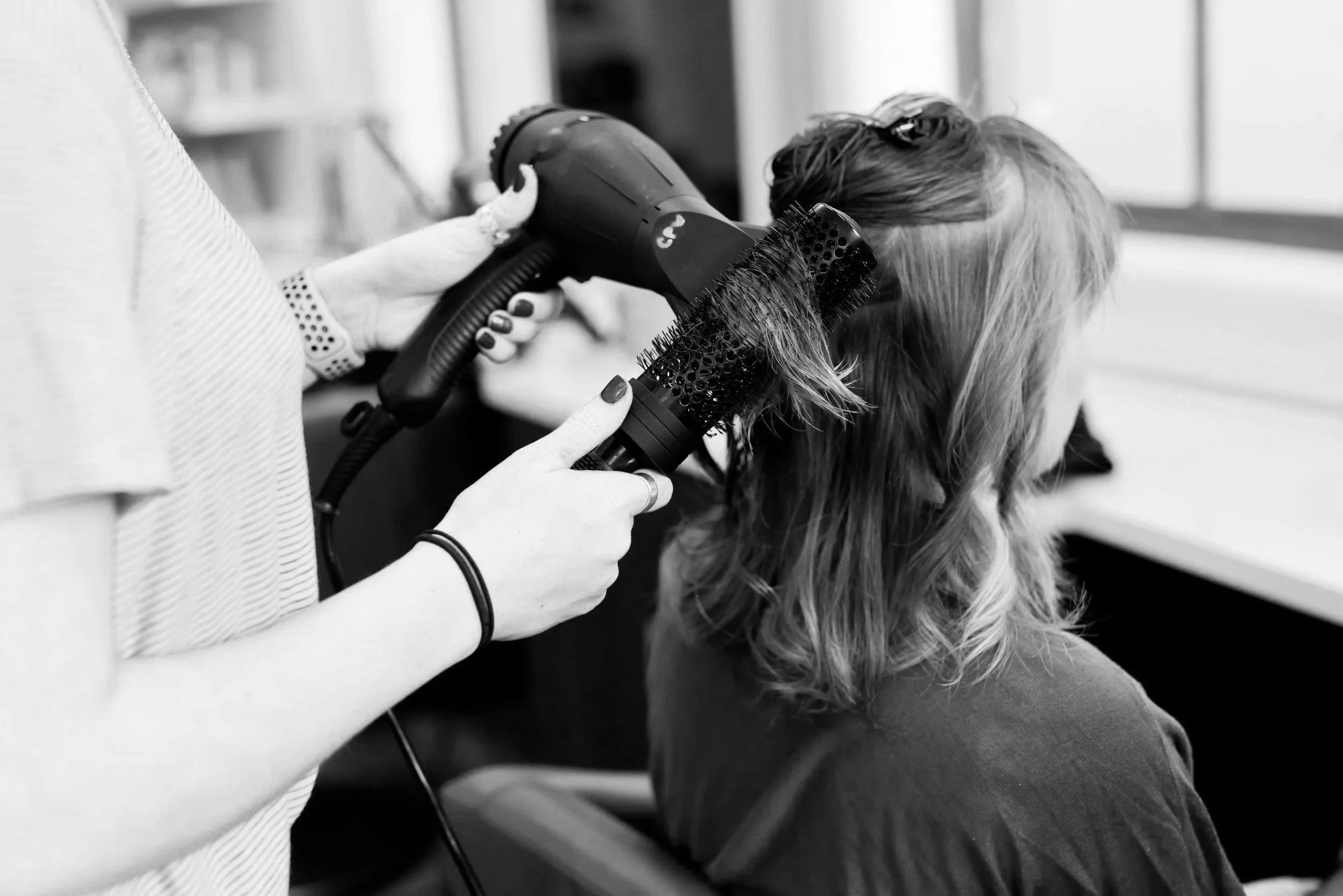 A person drying or styling a woman's hair using a handheld hair dryer and a round brush. NHR Studios | Nashville Hair Replacement | J. Bangs Salon