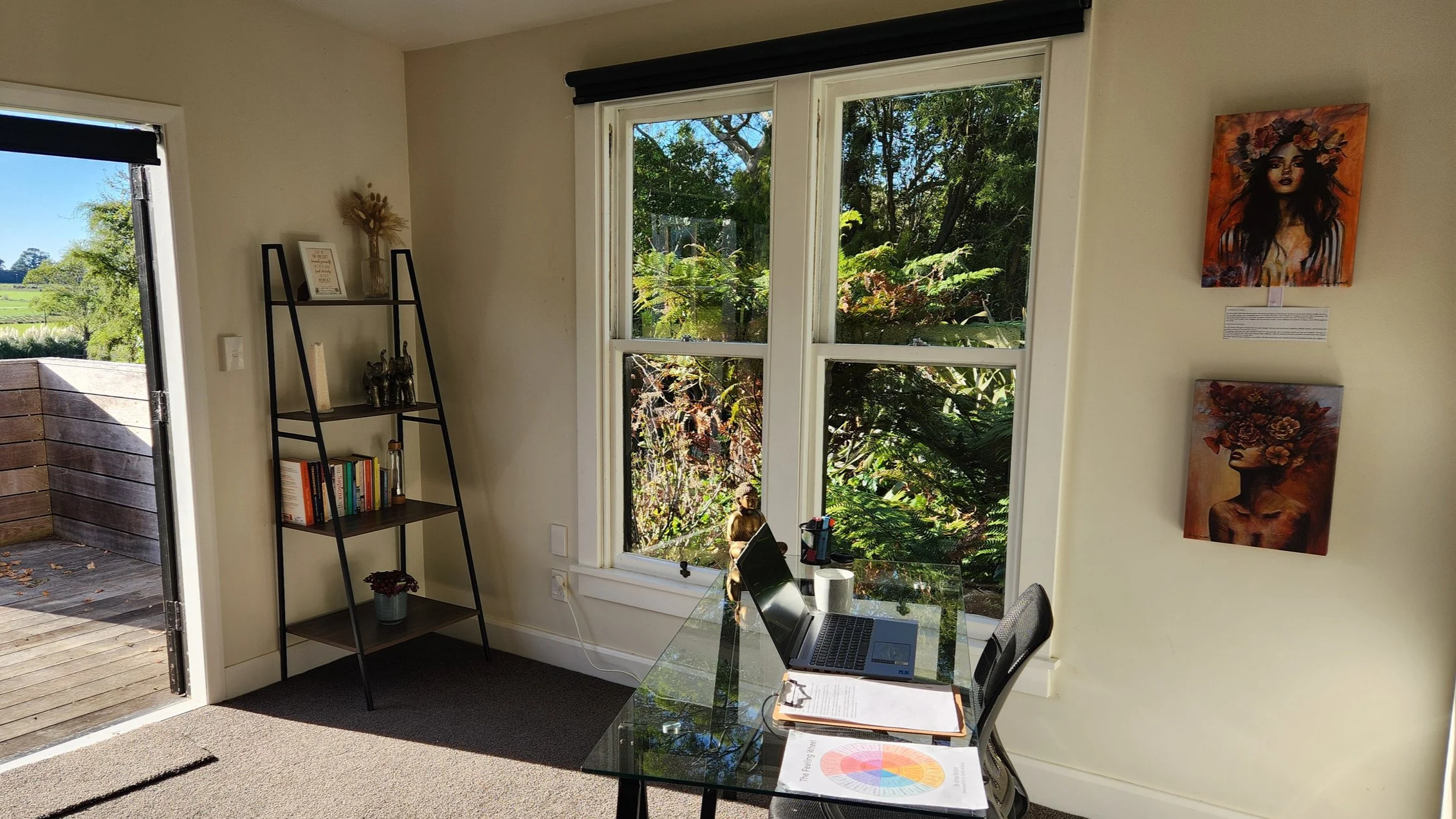 Interior of a room with a large front window showing lush green trees outside. The room has a computer desk with an open laptop, papers, and a chair. A black metal bookshelf with books and decorative items is next to the door leading outside. There are paintings on the wall, including a portrait of a woman with a floral headpiece.