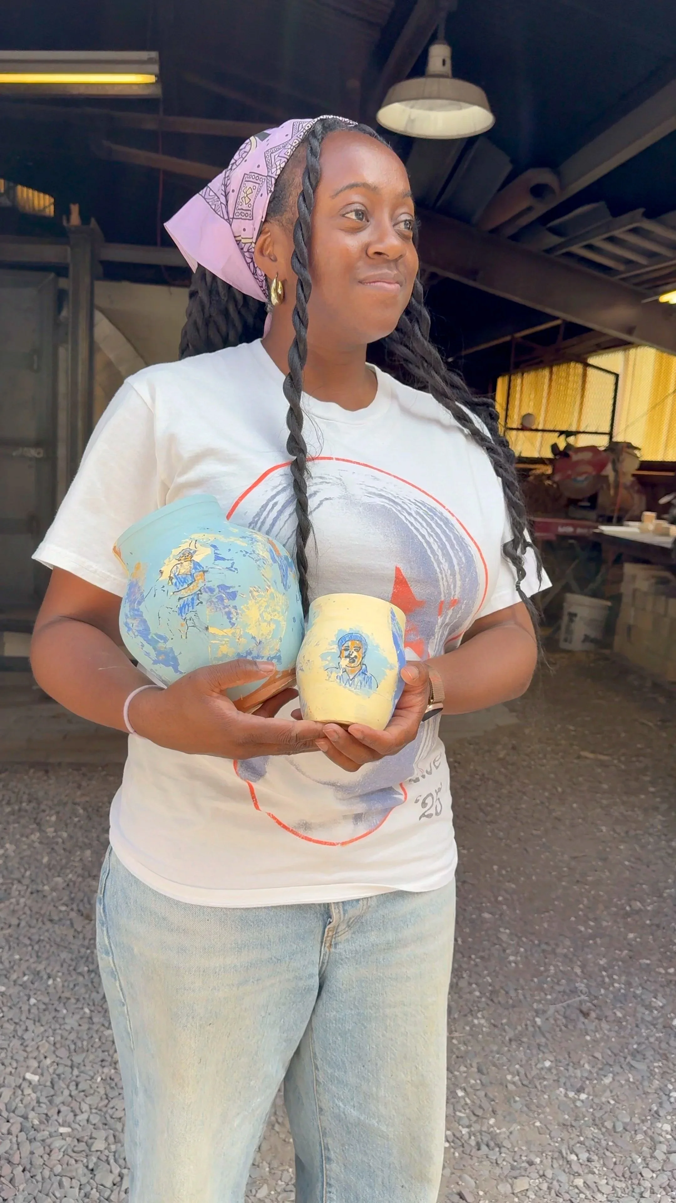 Woman with long braided hair, wearing a pink bandana, holding two painted pottery pieces with portraits, standing in a workshop or warehouse.